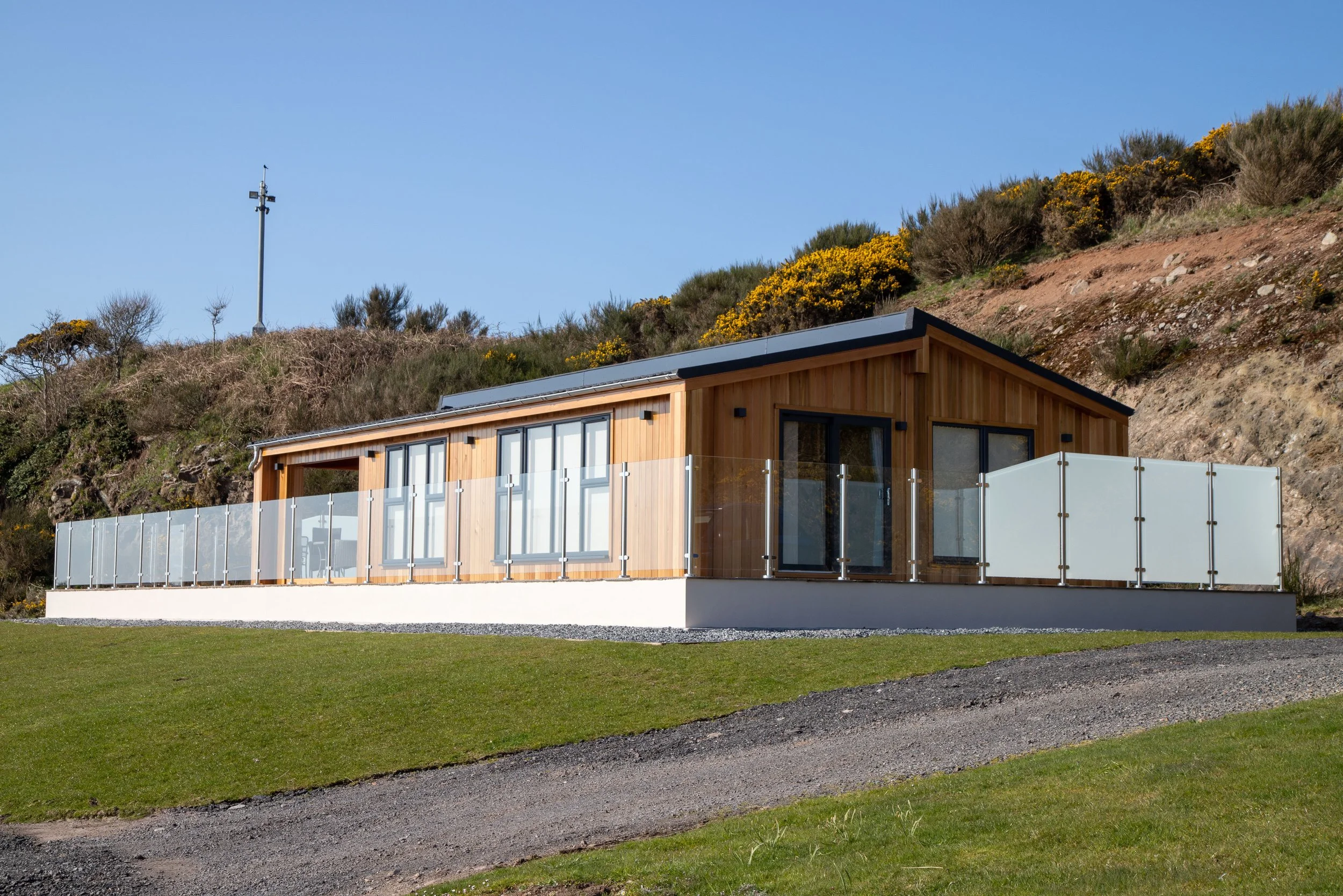 Modern house with wooden exterior and glass railings on a grassy hill, with a hillside and blue sky in the background.