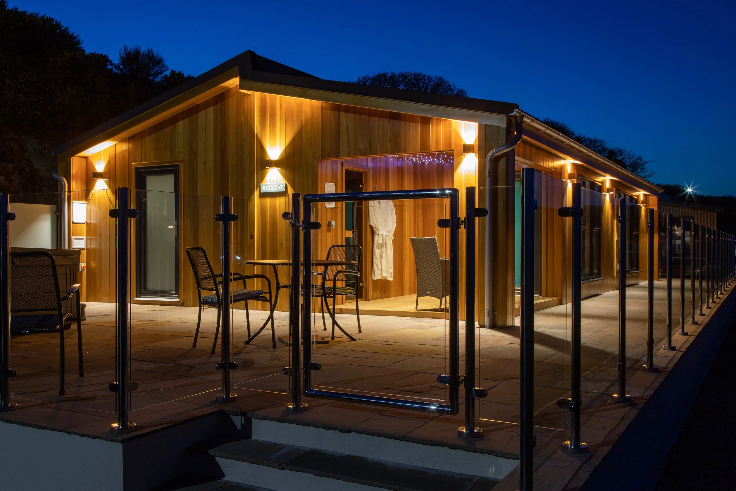 Night view of a modern house with wooden exterior and a spacious outdoor patio, illuminated with warm lights, featuring patio furniture and glass railings.
