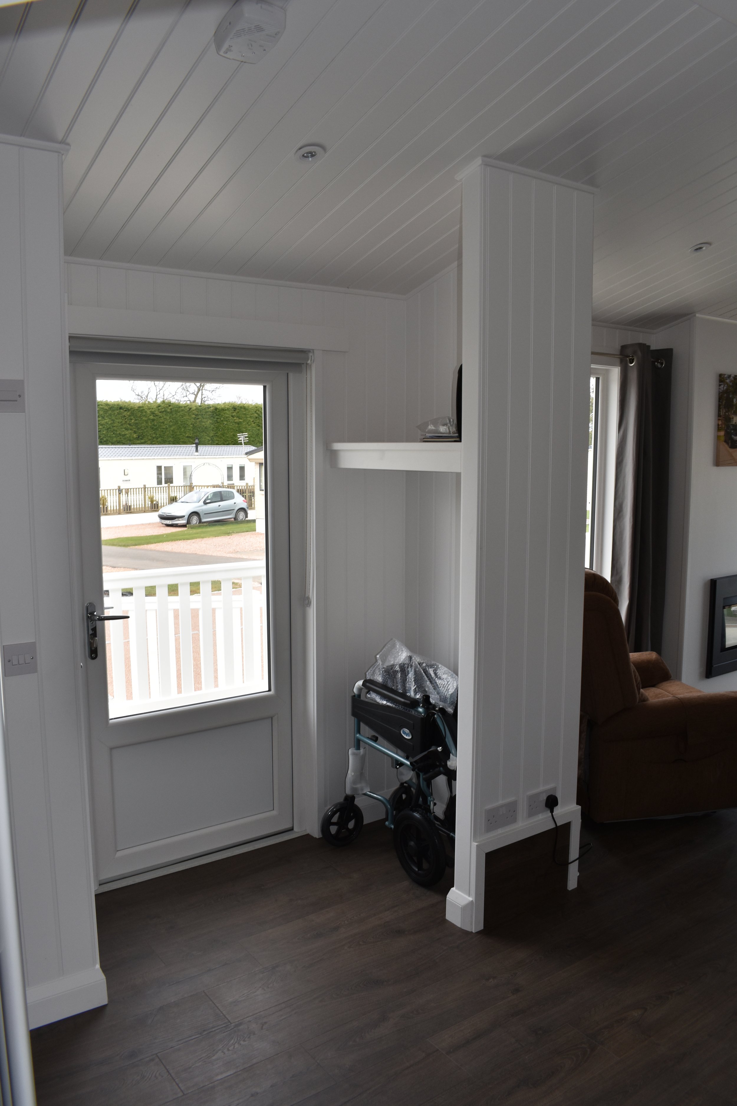 A view of a home's interior entryway showing a glass door leading outside. A walker with black wheels is positioned against a white paneled wall. A brown couch and a fireplace are visible in the living room to the right.