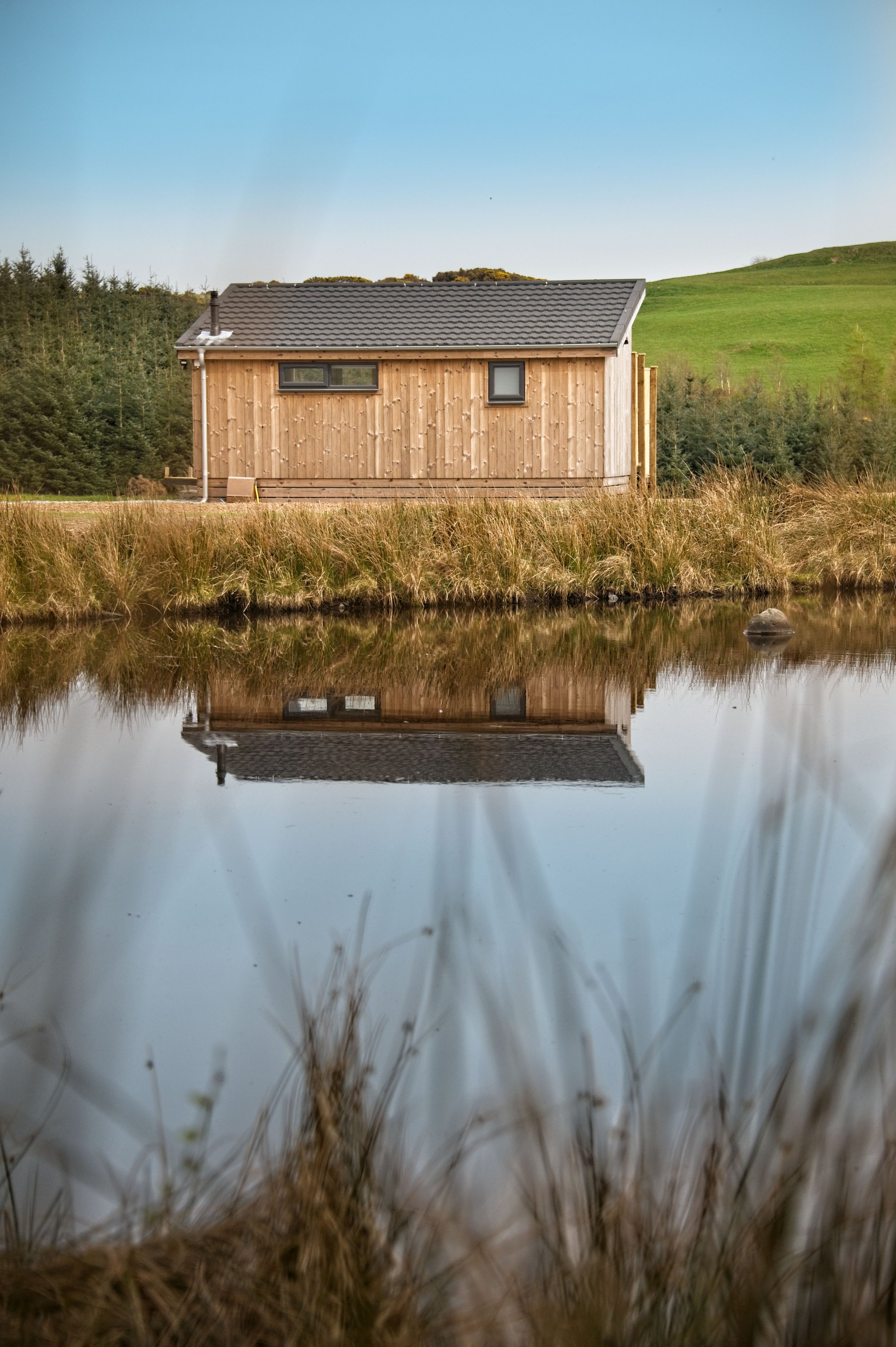 A wooden house with a gray roof reflected in a calm body of water, with grassy banks, trees, and a hill in the background under a blue sky.