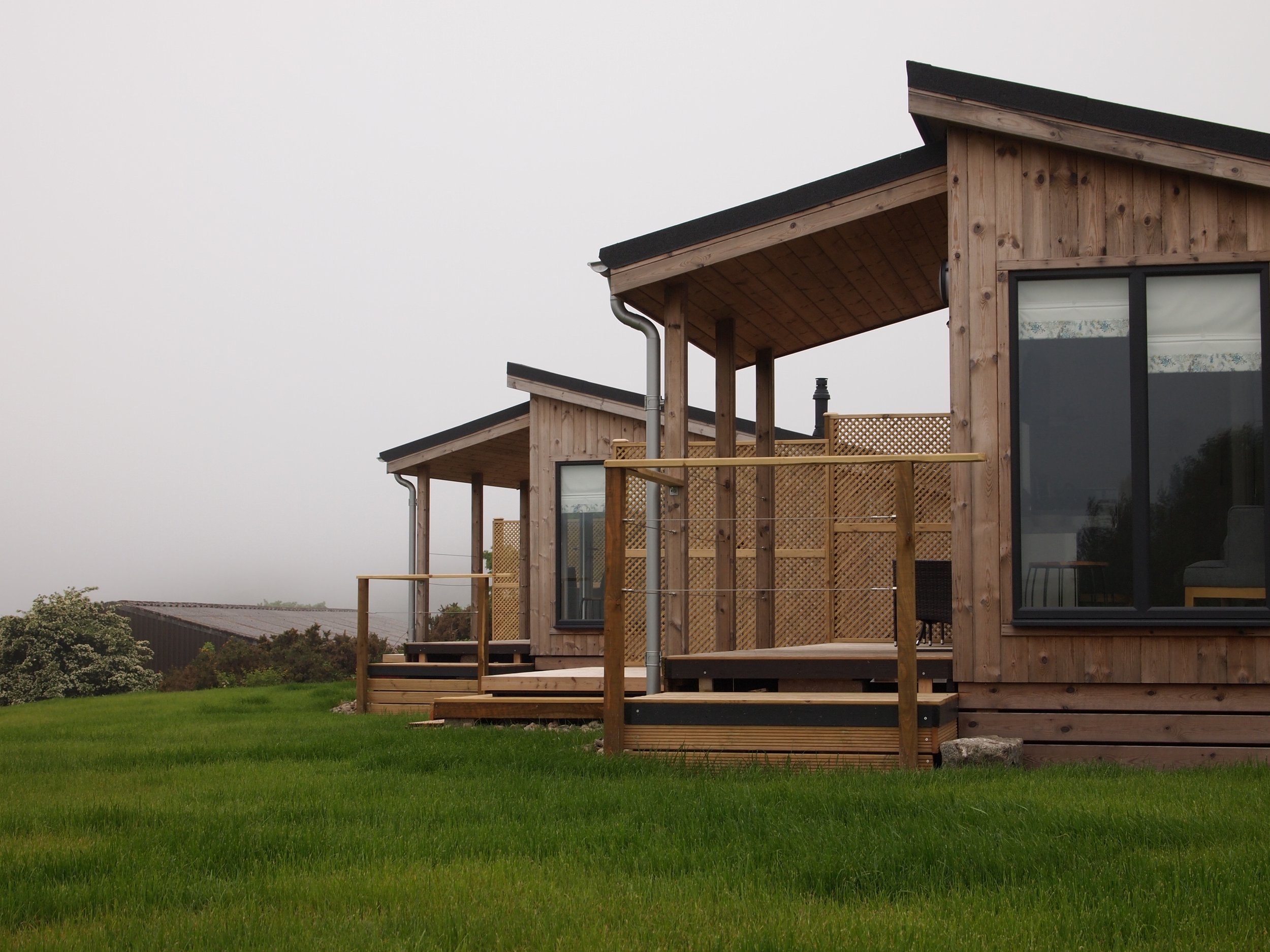 Two modern wooden houses with large glass windows and decks, surrounded by green grass and overcast sky.