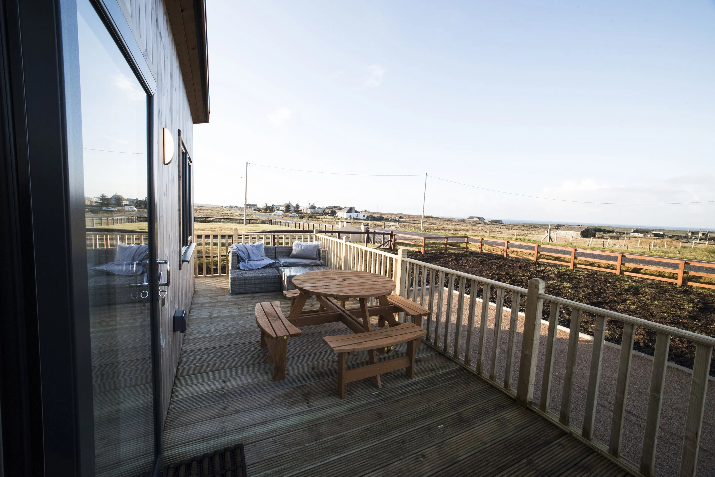 A wooden deck with outdoor furniture, including a picnic table and a wicker seating area with cushions, overlooking open fields with fences and distant buildings under a clear sky.