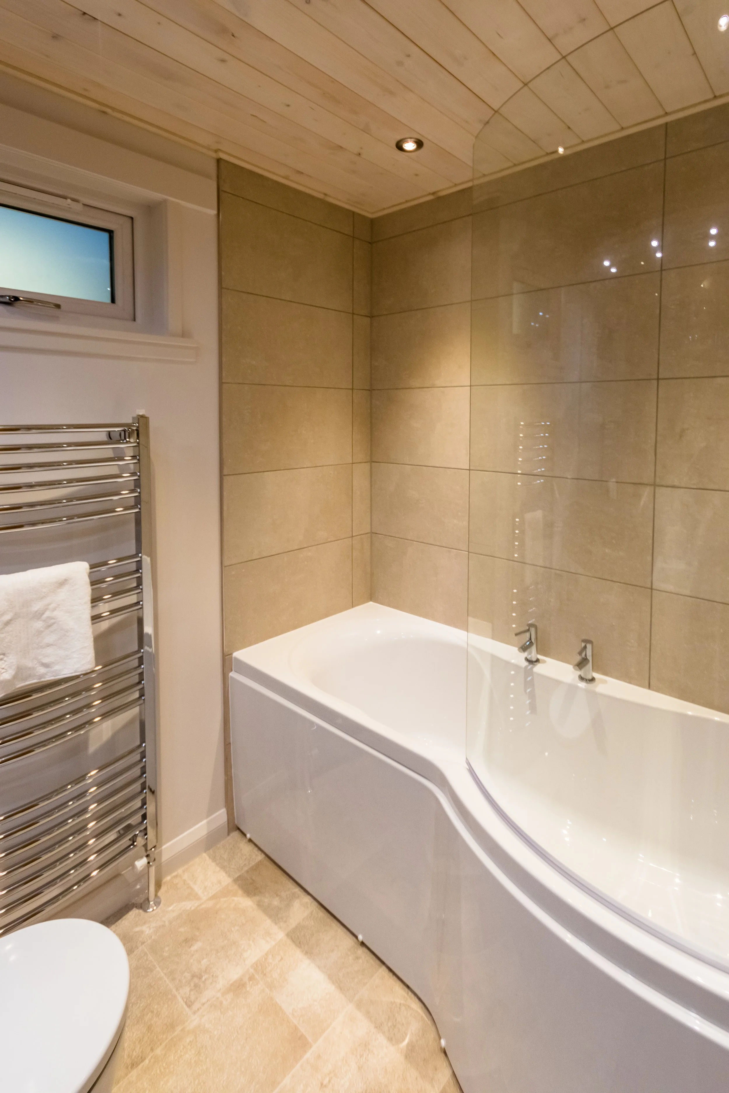 Bathroom with a corner bathtub, beige tiled walls, wooden ceiling, and a chrome towel heater with a white towel hanging on it. A small window is visible near the ceiling.