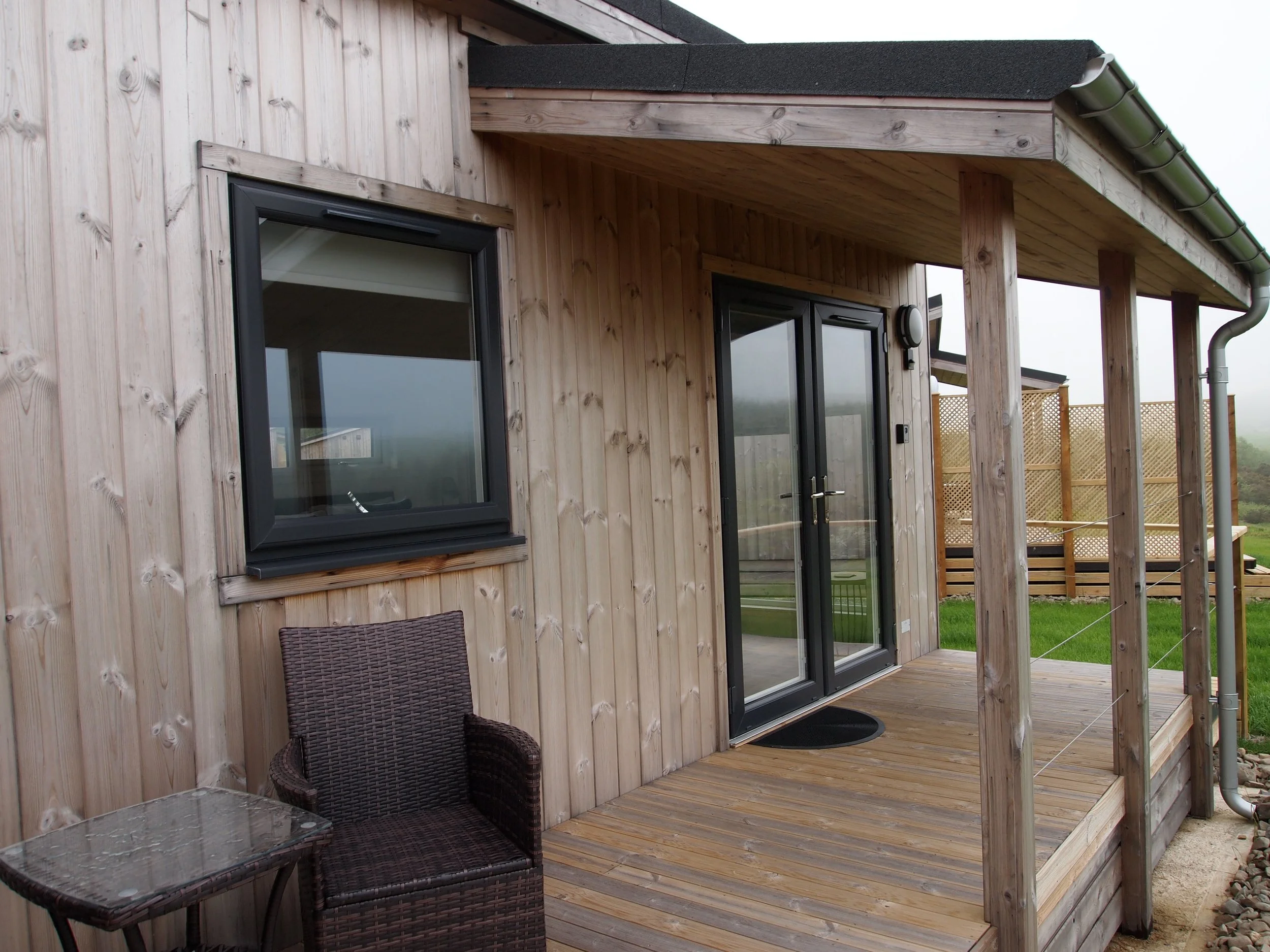 Wooden house exterior with sliding glass door, a window, and a small porch with patio furniture, set in a grassy yard.