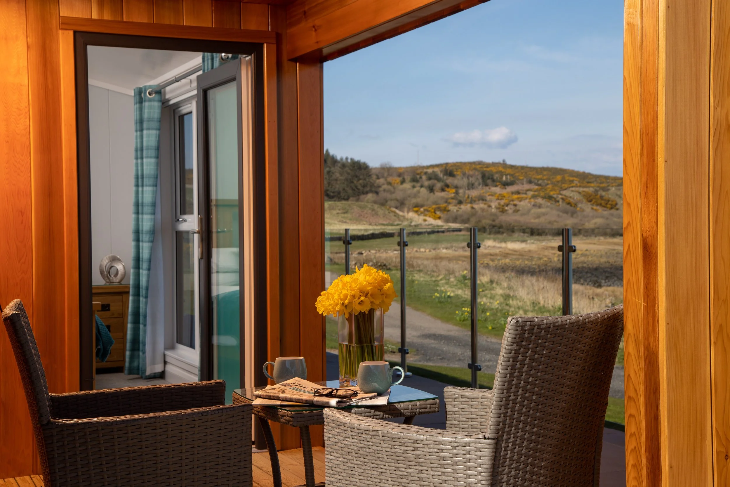 Interior view of a cozy wooden room with a small balcony. There is a table set with two mugs, sunglasses, newspapers, and a vase of yellow flowers. Outside, a scenic landscape with hills and a partly cloudy sky is visible.
