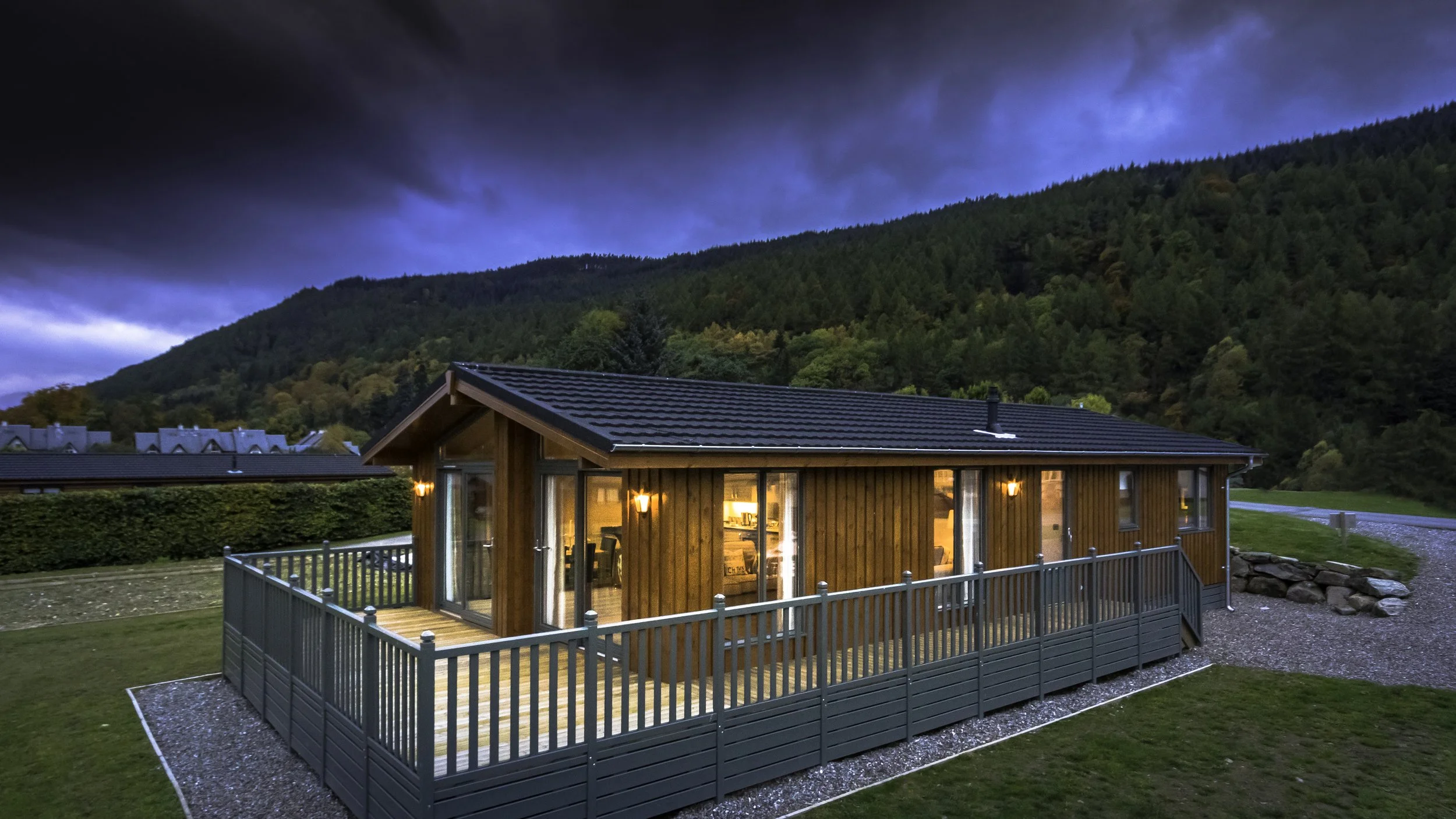 A wooden house with a deck, lit by interior lights, set against a backdrop of a green hillside and dark cloudy sky during dusk.