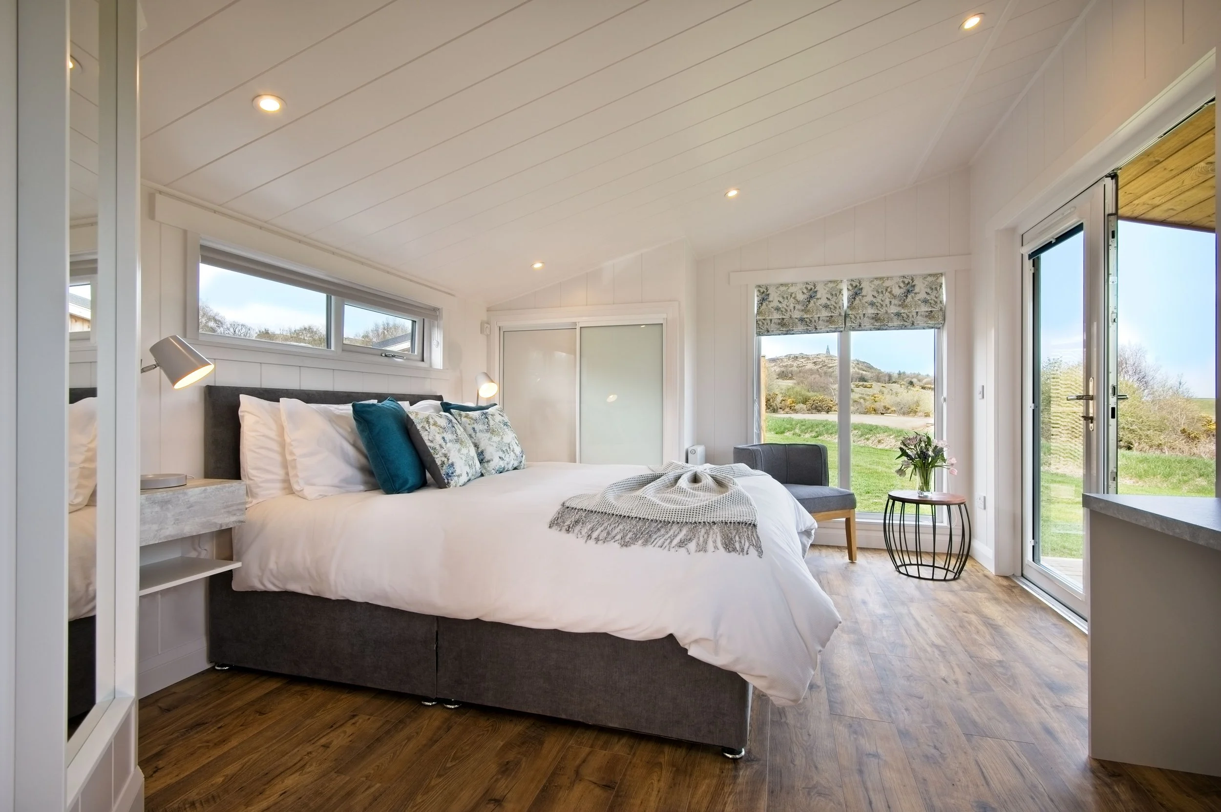 Bright bedroom with white walls and ceiling, large bed with white bedding and decorative pillows, wooden flooring, side tables with lamps, large windows with rolled shades, a chair with a small table holding flowers, and glass door leading outside to
