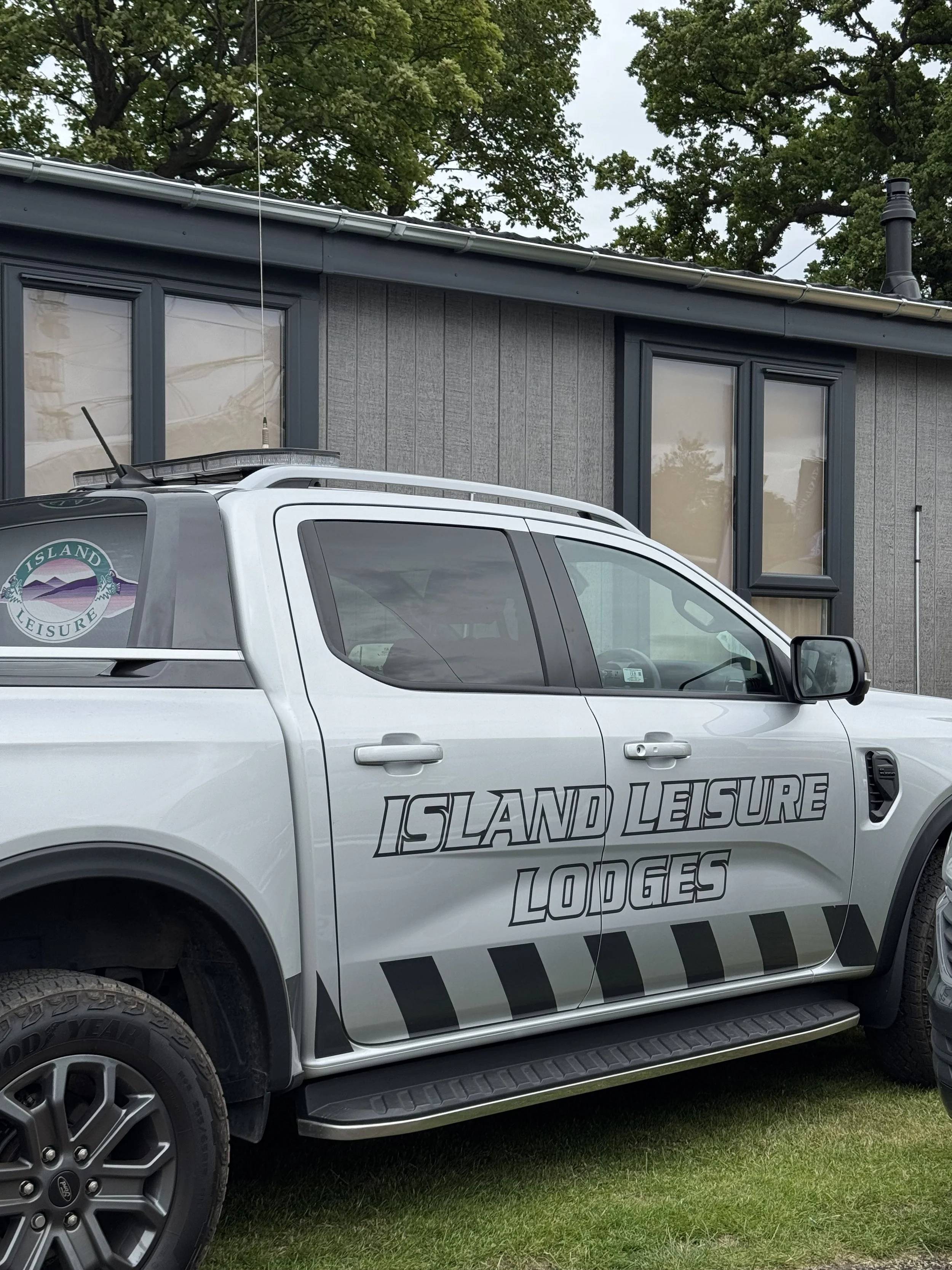 A white pickup truck with 'Island Leisure Lodges' branding parked on grass in front of a gray building with dark window frames and a tree with green leaves in the background.