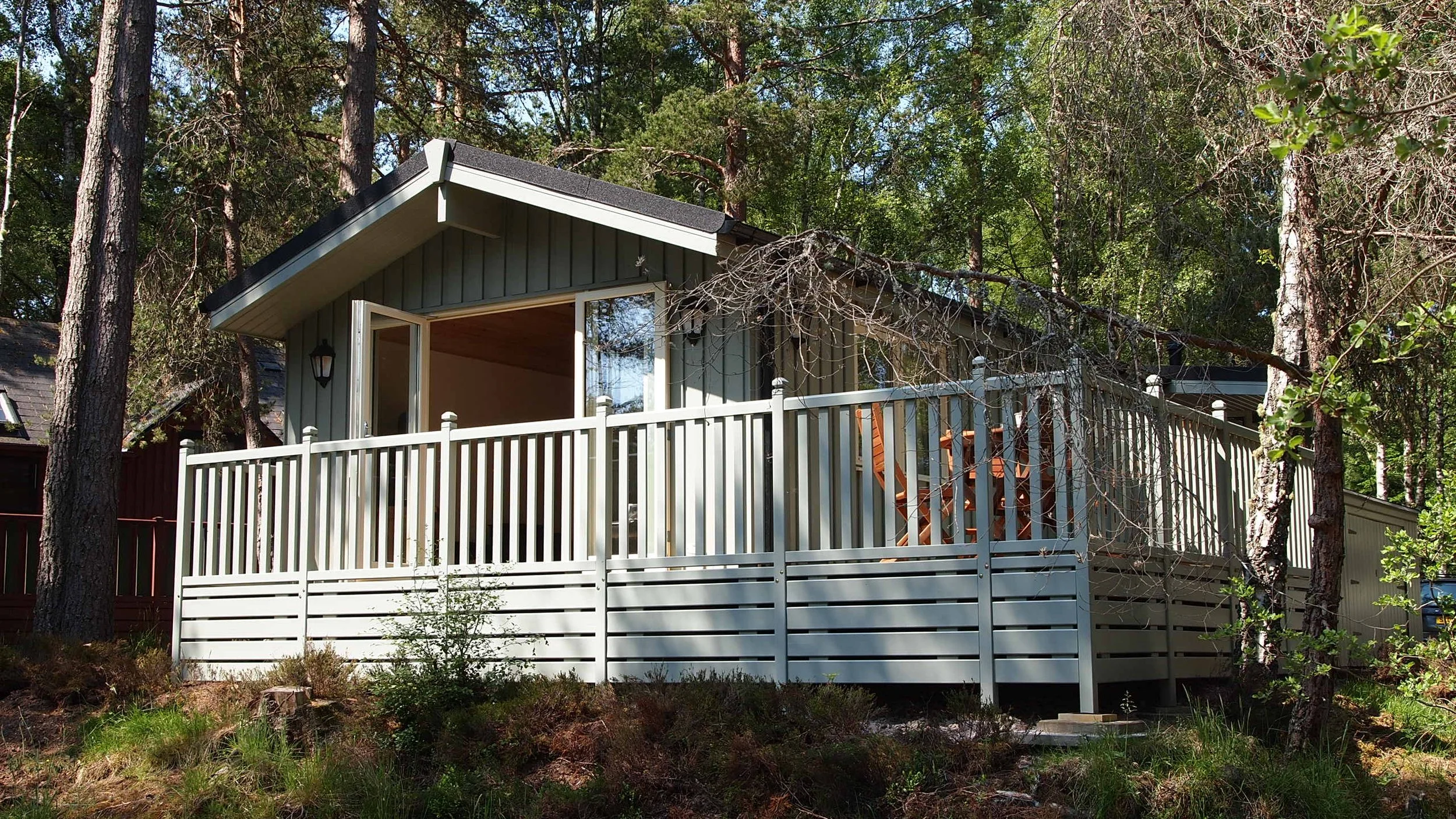 A house with a gray exterior and a wooden ceiling porch, surrounded by tall trees, with a white railing, and outdoor chairs on the deck.