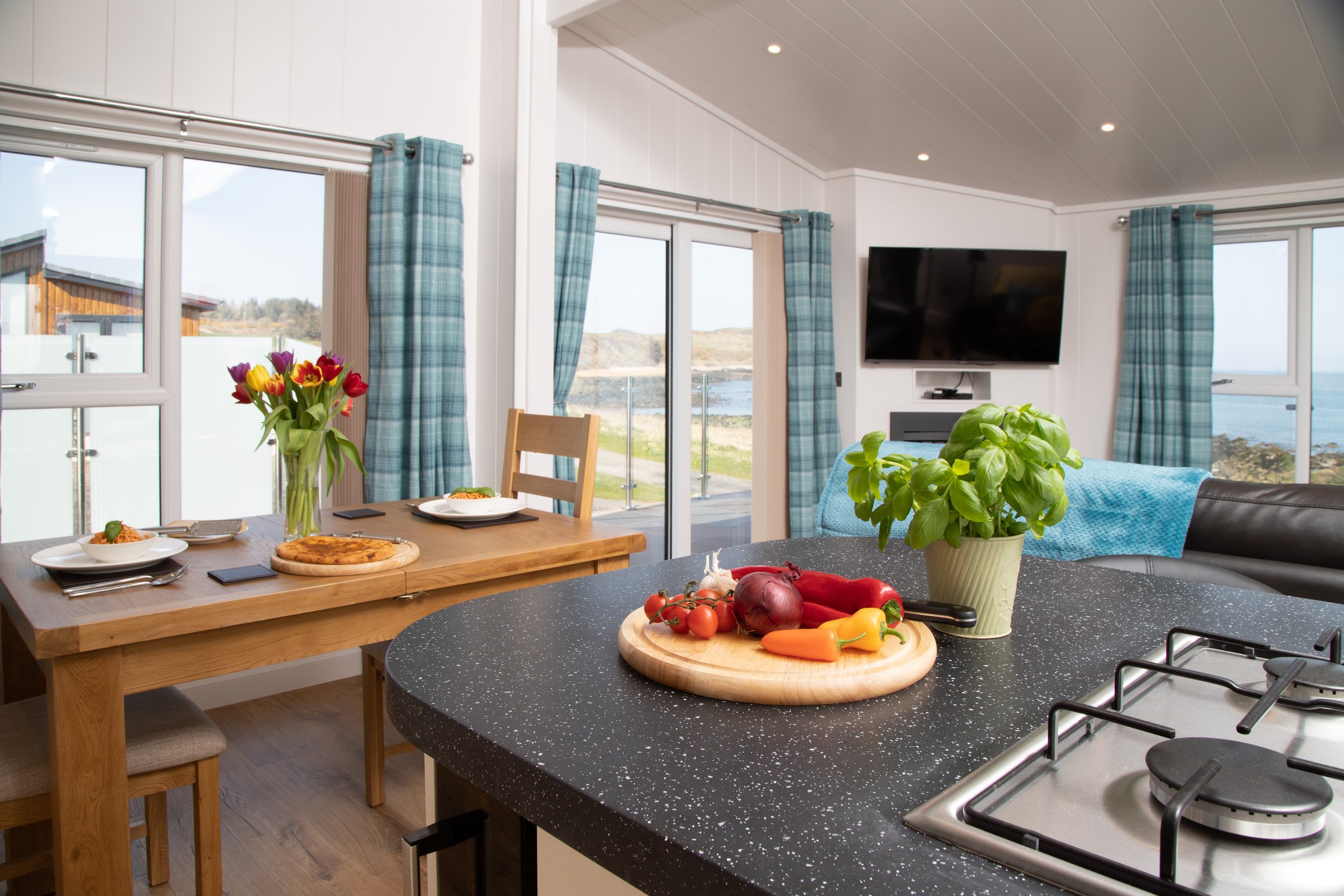 View of a bright, airy kitchen and dining area with large windows and a scenic outdoor view, a black granite kitchen island with a basil plant and fresh vegetables, a wooden dining table with plates, cutlery, and a vase of tulips.