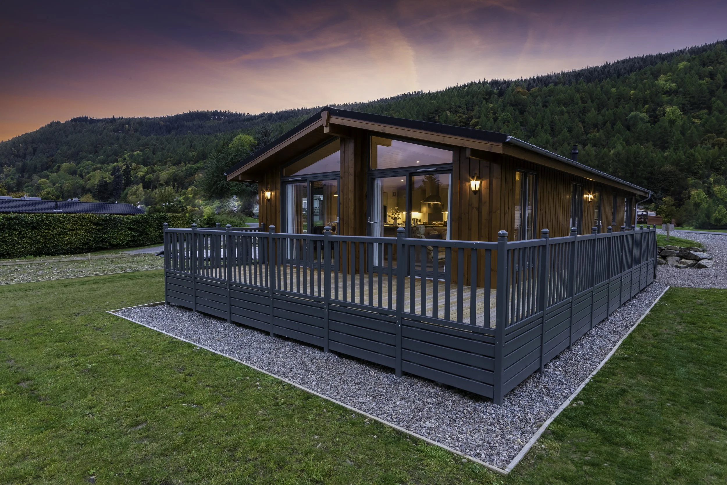 A modern wooden house with large glass windows and a balcony, surrounded by green grass and hills at sunset.