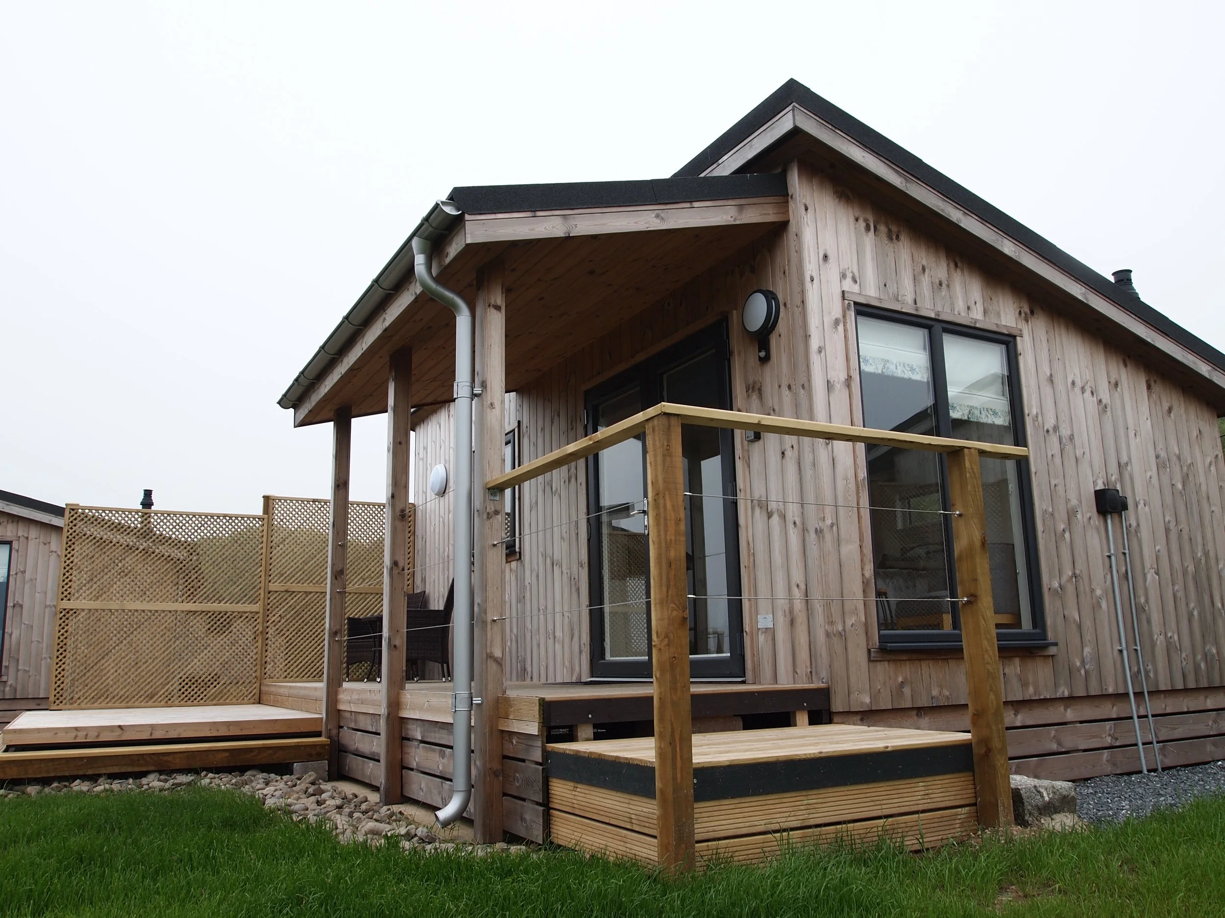 Wooden house with sliding glass doors and a small deck with railing, situated on a grassy lawn with a cloudy sky.
