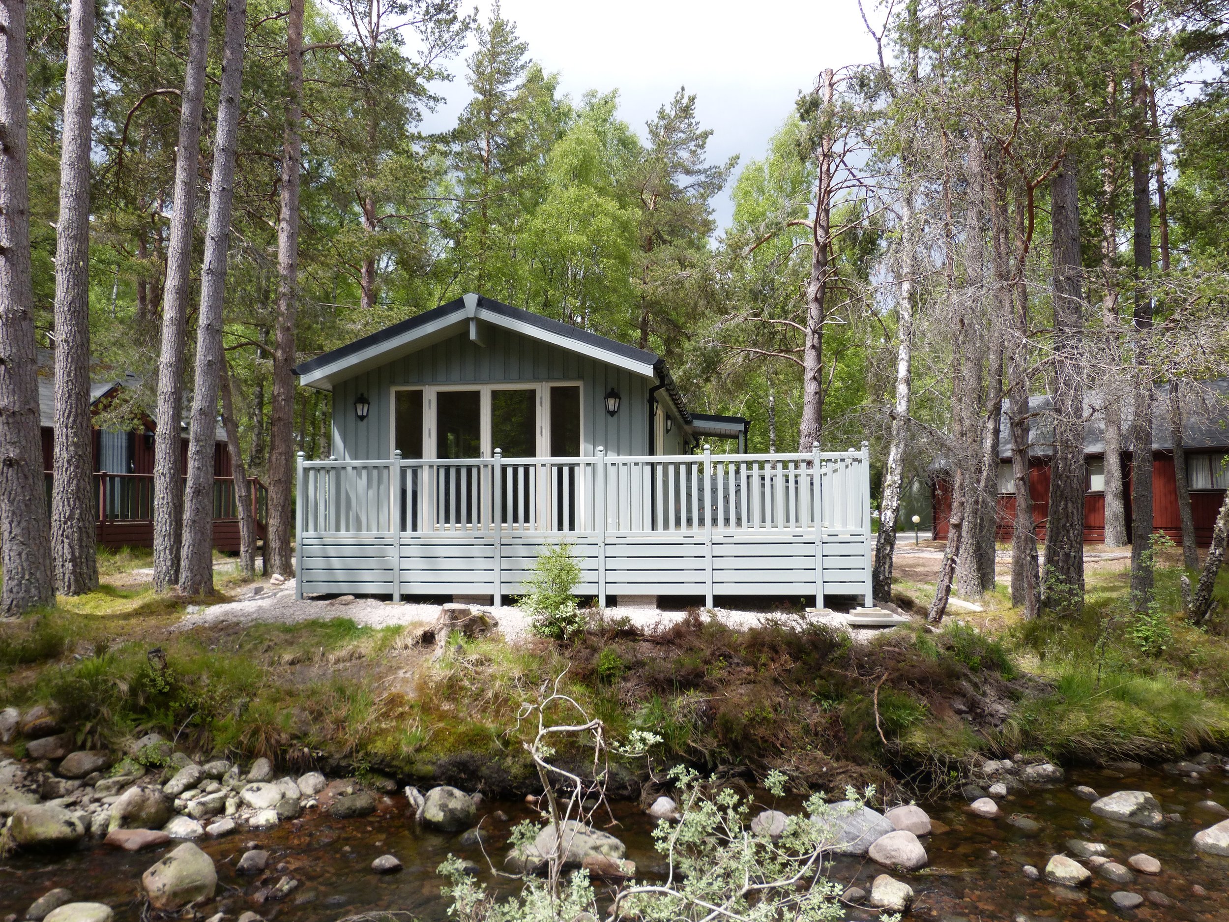 A small house with a gray exterior, a porch, and a black gable roof in a wooded area, with tall trees and a creek in the foreground.