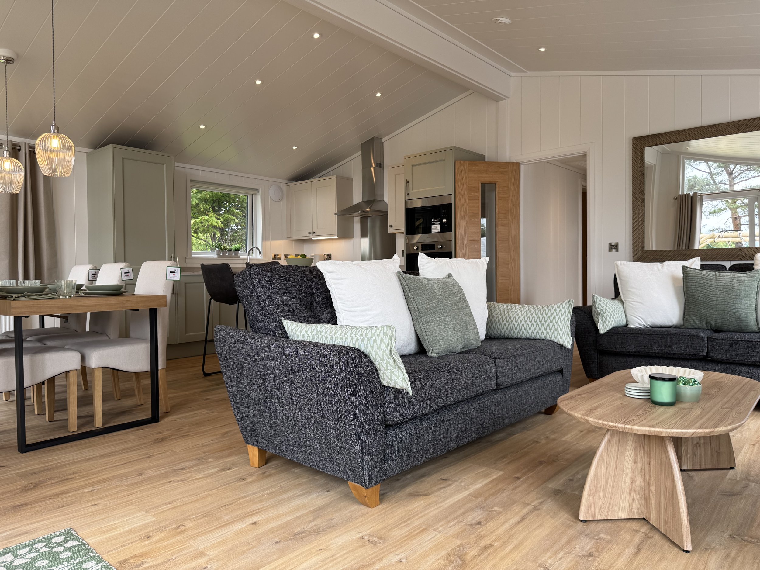Open-plan living room and kitchen with gray sofas, light wood coffee table, dining area, and kitchen cabinets, with a window showing greenery outside.