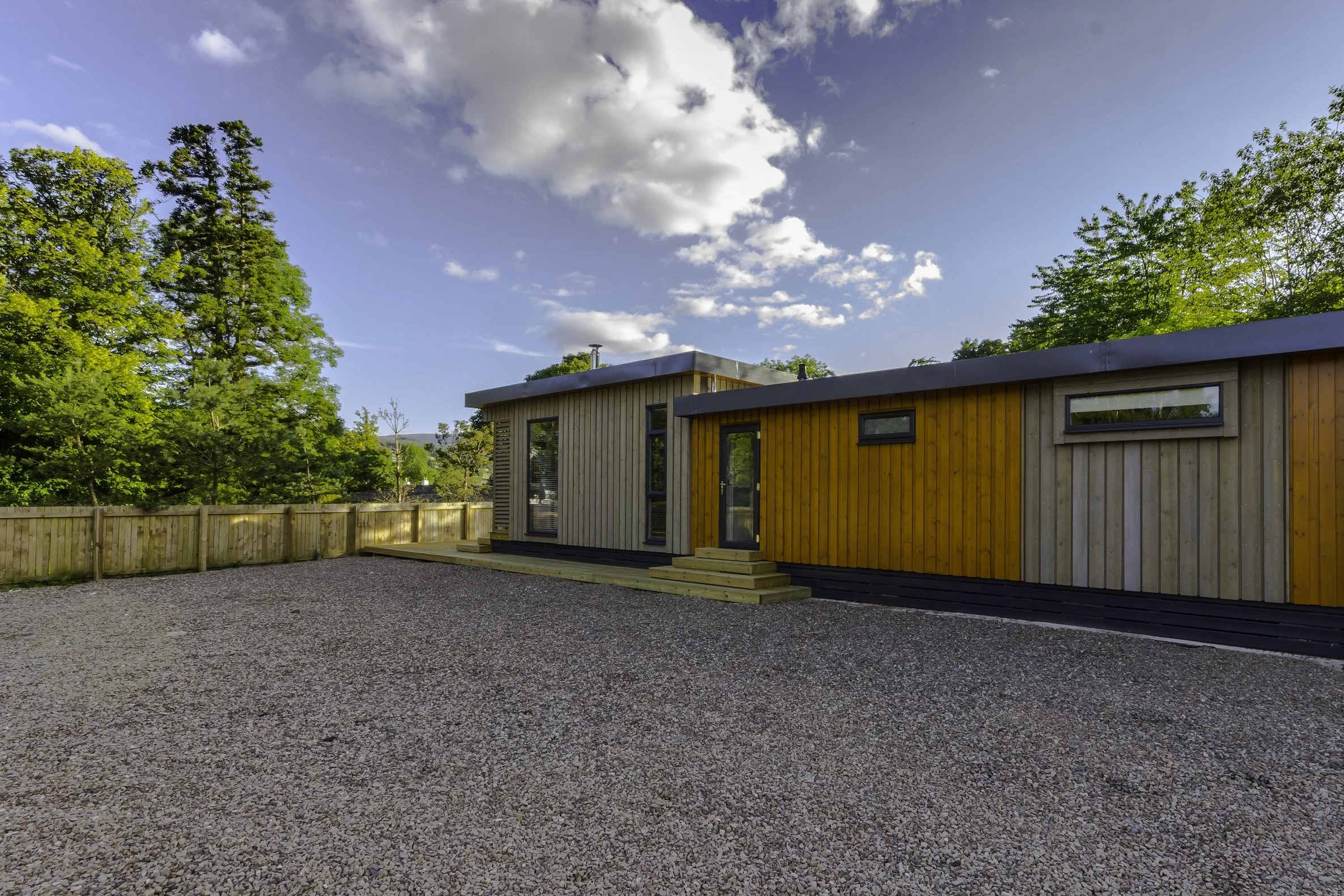 A modern single-story house with custom wooden exterior walls, surrounded by a gravel yard, with a wooden fence and green trees in the background, under a partly cloudy sky.