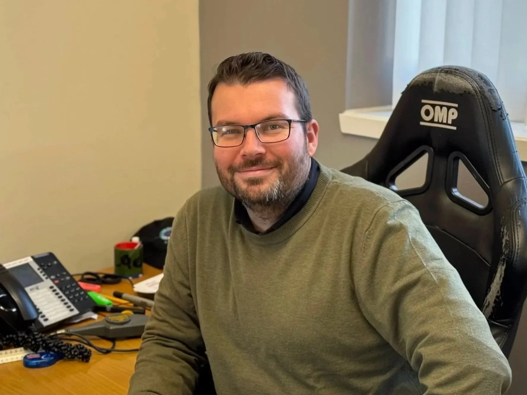 A man with glasses and a beard sitting at an office desk with a smile. The desk has a phone, tape, and miscellaneous office supplies. He is sitting in a black OMP racing-style chair near a window with blinds.