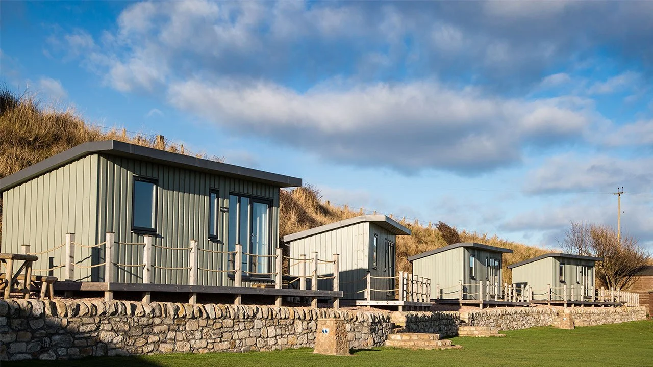 Four small, green, metal cabins on a stone foundation with a grassy area in front, under a partly cloudy sky.