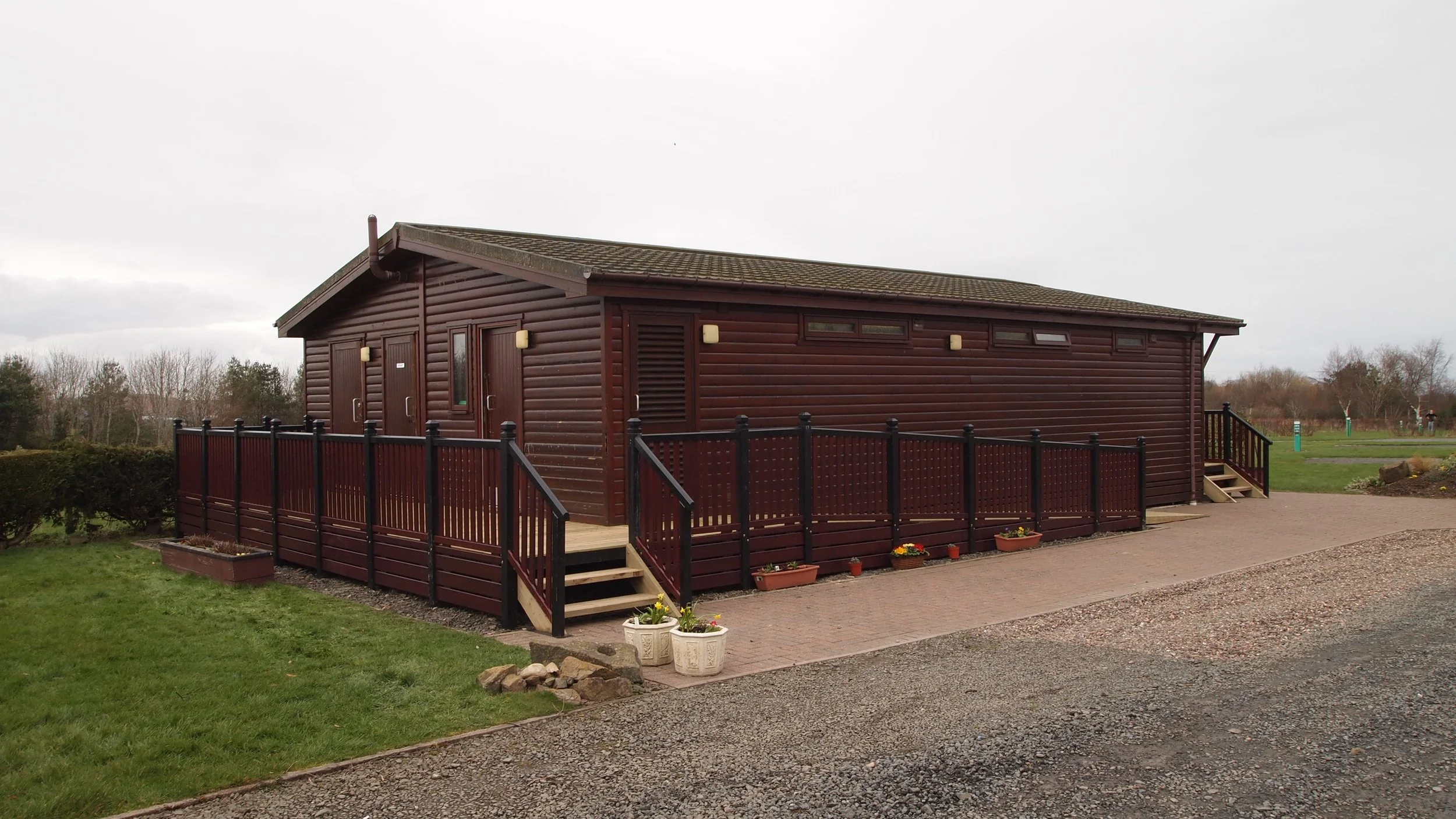 A single-story wooden building with a ramp and stairs, surrounded by grass and a paved walkway, with trees and a cloudy sky in the background.