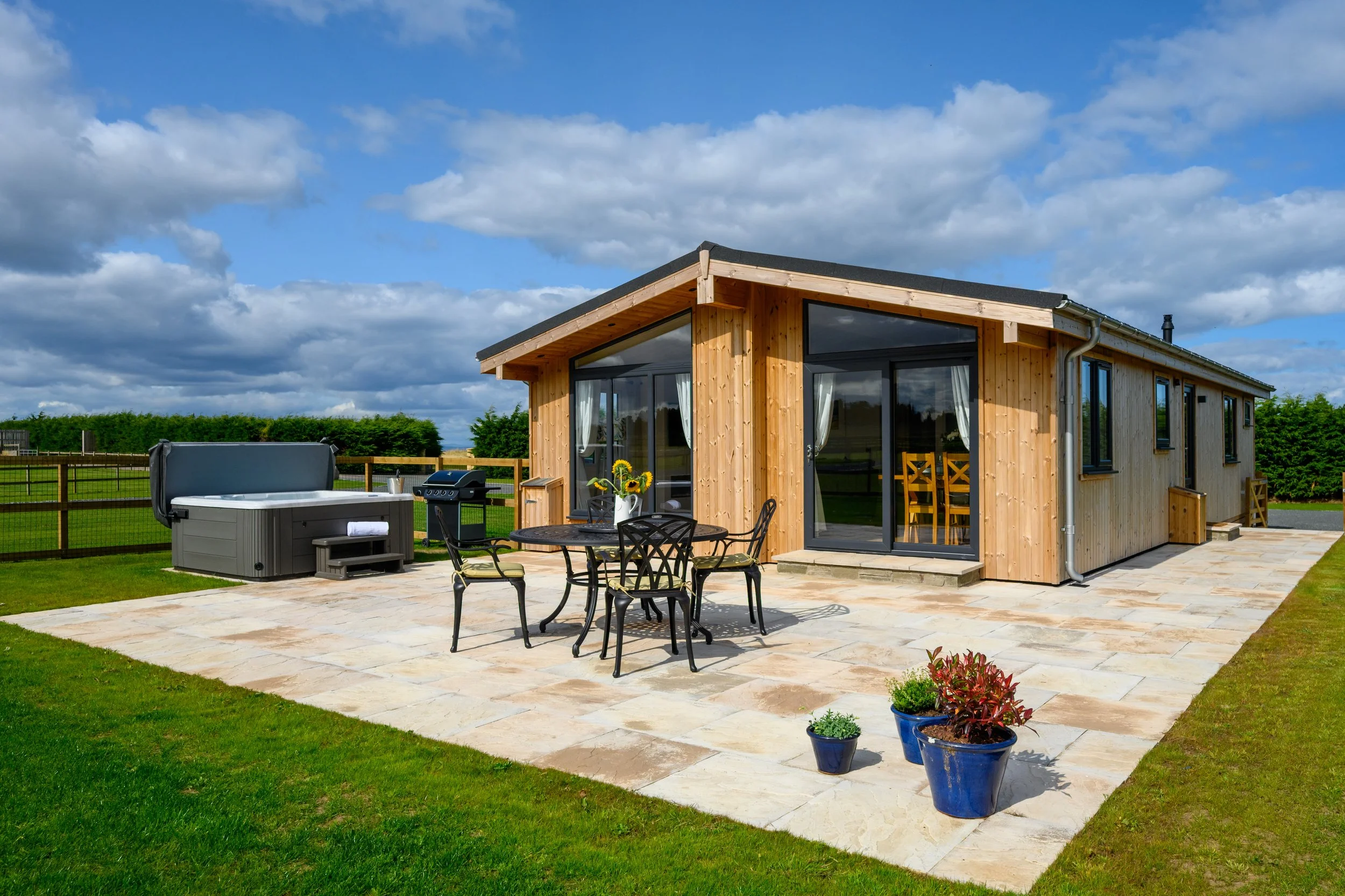 A modern wooden house with large glass doors and windows, a patio with a round table and four chairs, a hot tub on the left, potted plants, and a grassy yard under a partly cloudy sky.