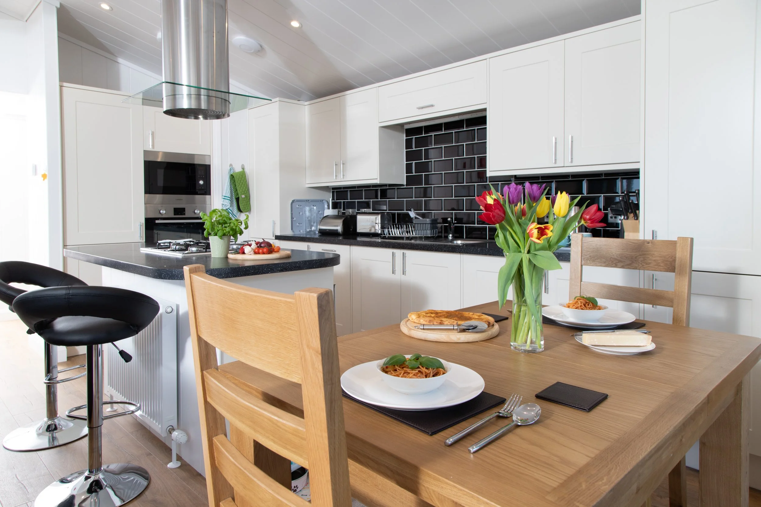 Kitchen with white cabinets, black backsplash, and a wooden dining table. The table has plates of spaghetti, a pizza, a vase of colorful tulips, and cutlery. There are two black barstools at the counter.