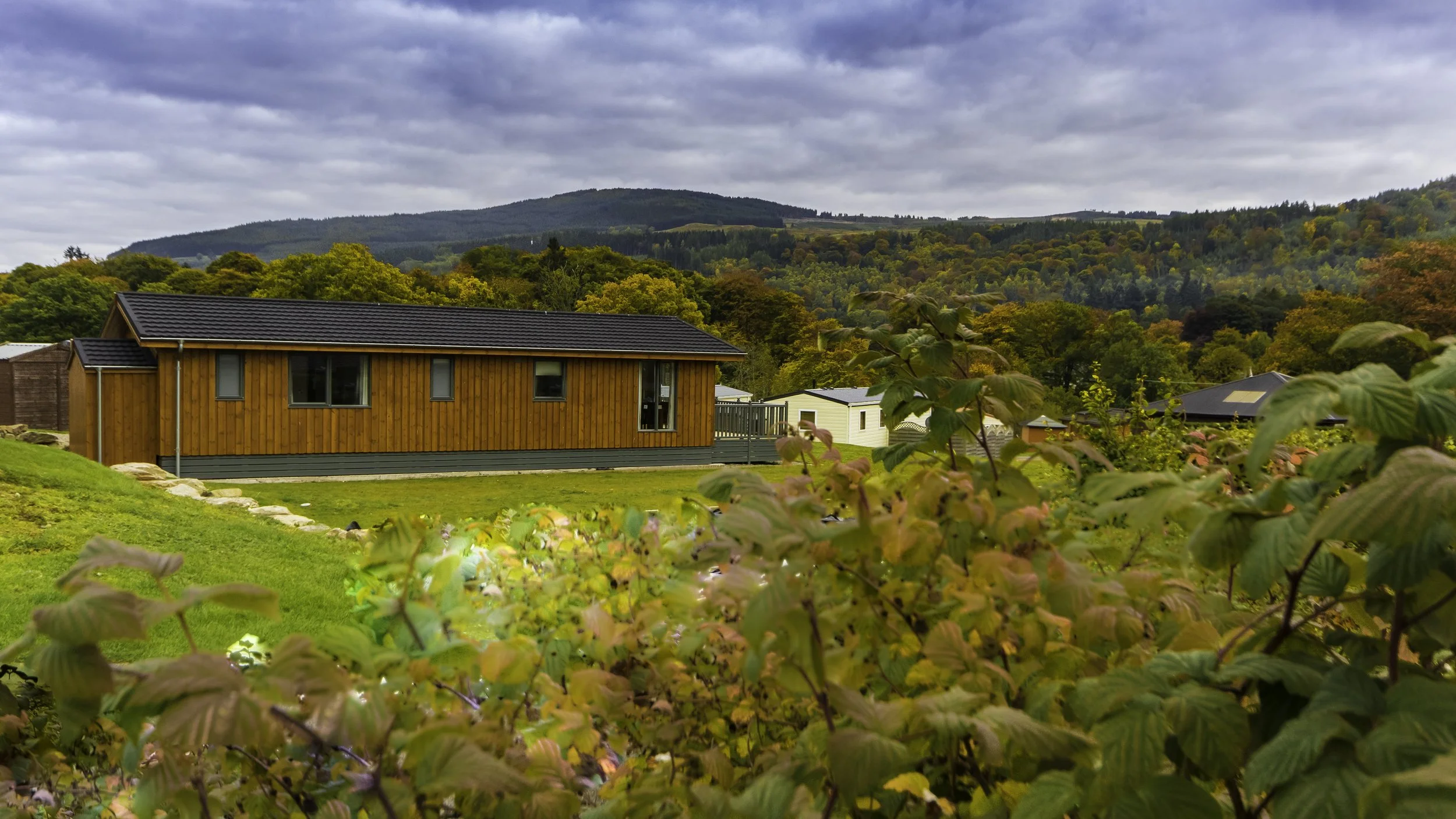 A modern wooden house with a black roof on a grassy hill, surrounded by autumn-colored trees and hills in the background.