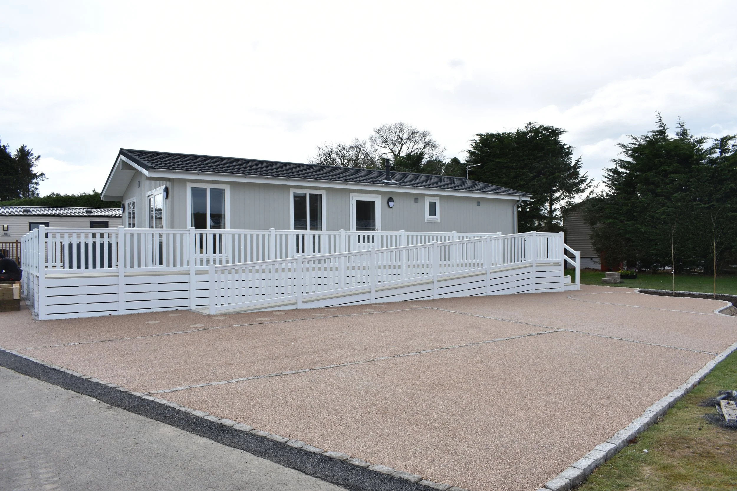 A light gray house with a dark roof surrounded by a white fence and a gravel driveway, with trees in the background.