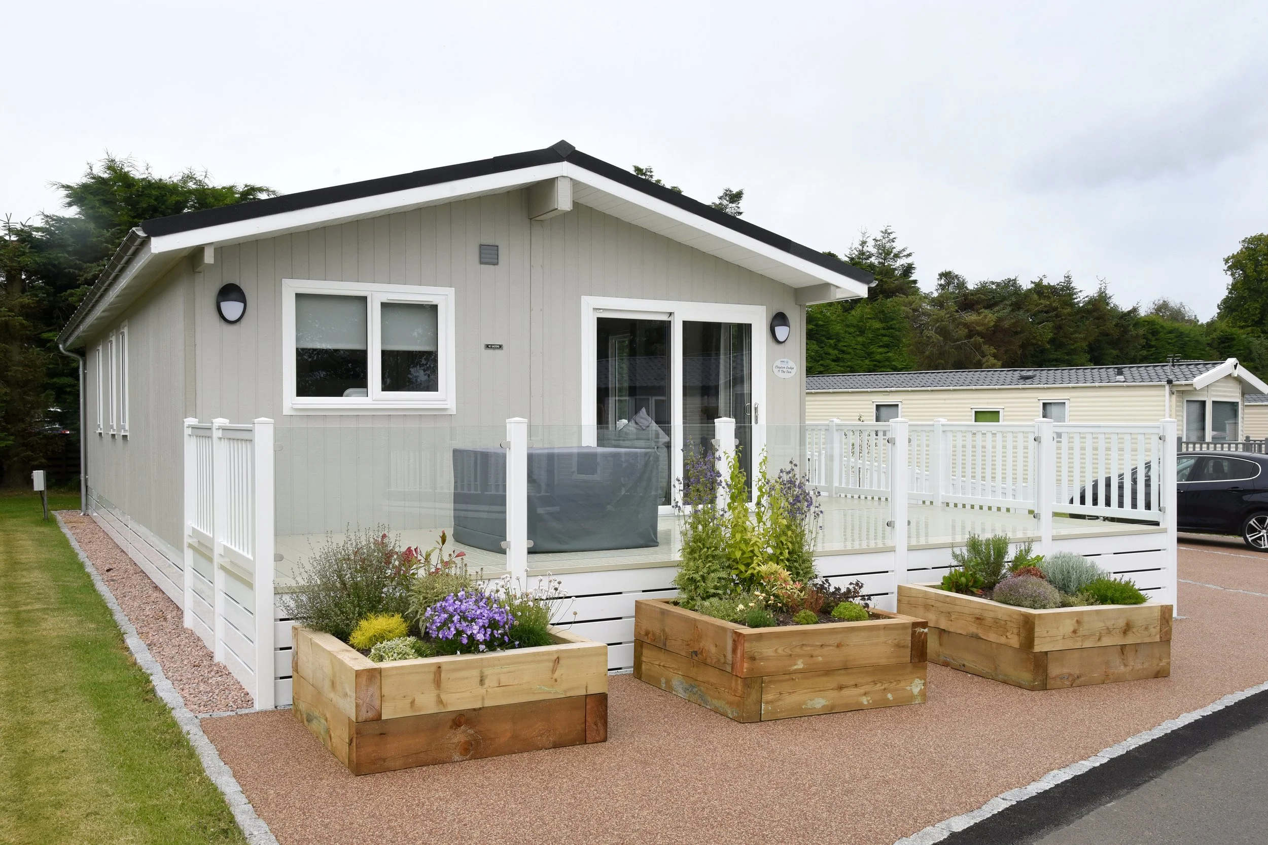 A small, modern house with a balcony, white railing, and garden planters, set against a backdrop of trees and a cloudy sky.