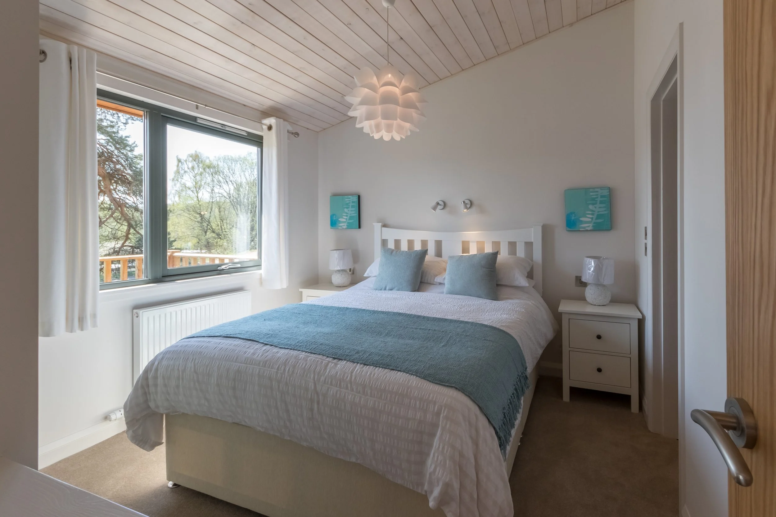 A bedroom with a large window overlooking trees, a white bed with blue pillows and a blue throw blanket, two white nightstands with lamps, wall art, and a white ceiling light fixture.