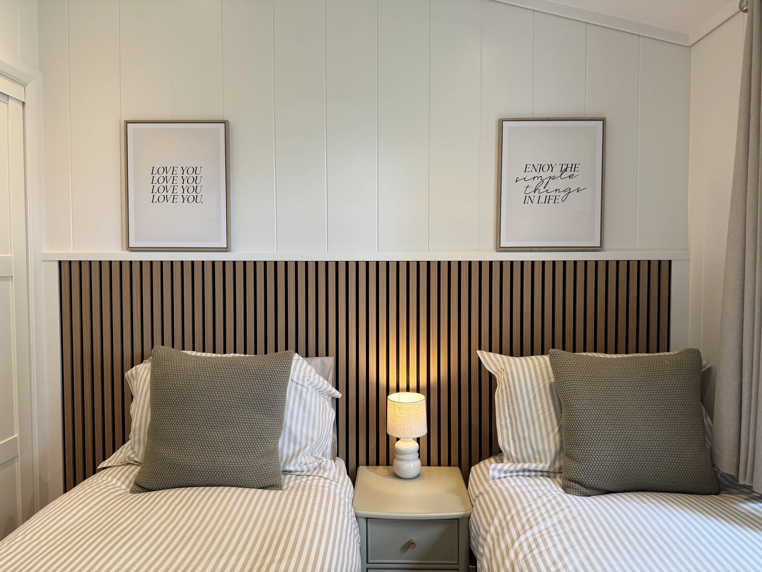 Two twin beds with striped bedding and large gray pillows, separated by a nightstand with a lamp, brown and black striped headboard, and two framed wall art quotes above the headboard.