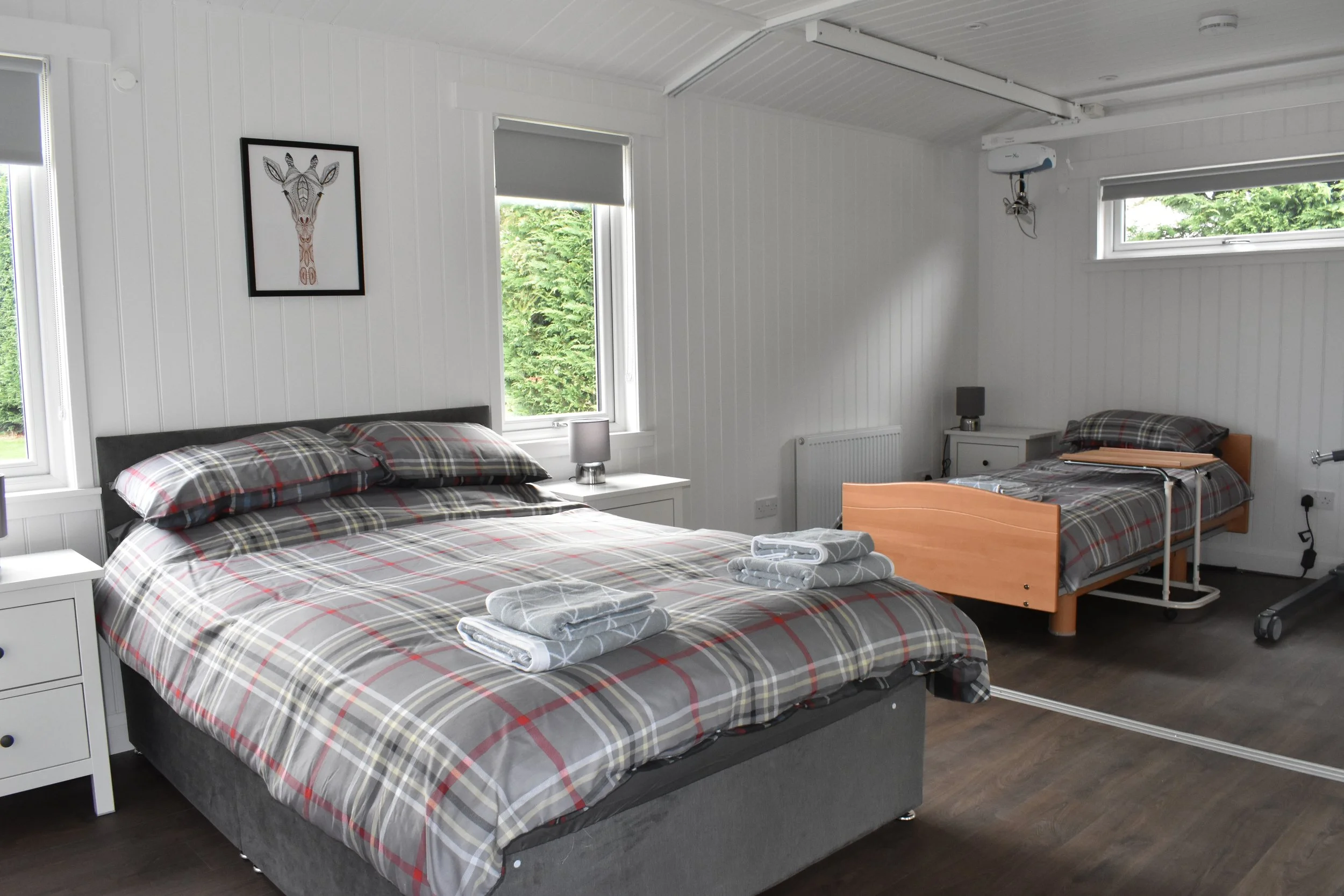 A room with a large bed covered in plaid bedding, and a smaller bed with editing support equipment. There are two nightstands with lamps, and a framed giraffe artwork on the wall. The room has white paneled walls, dark flooring, and windows with blin