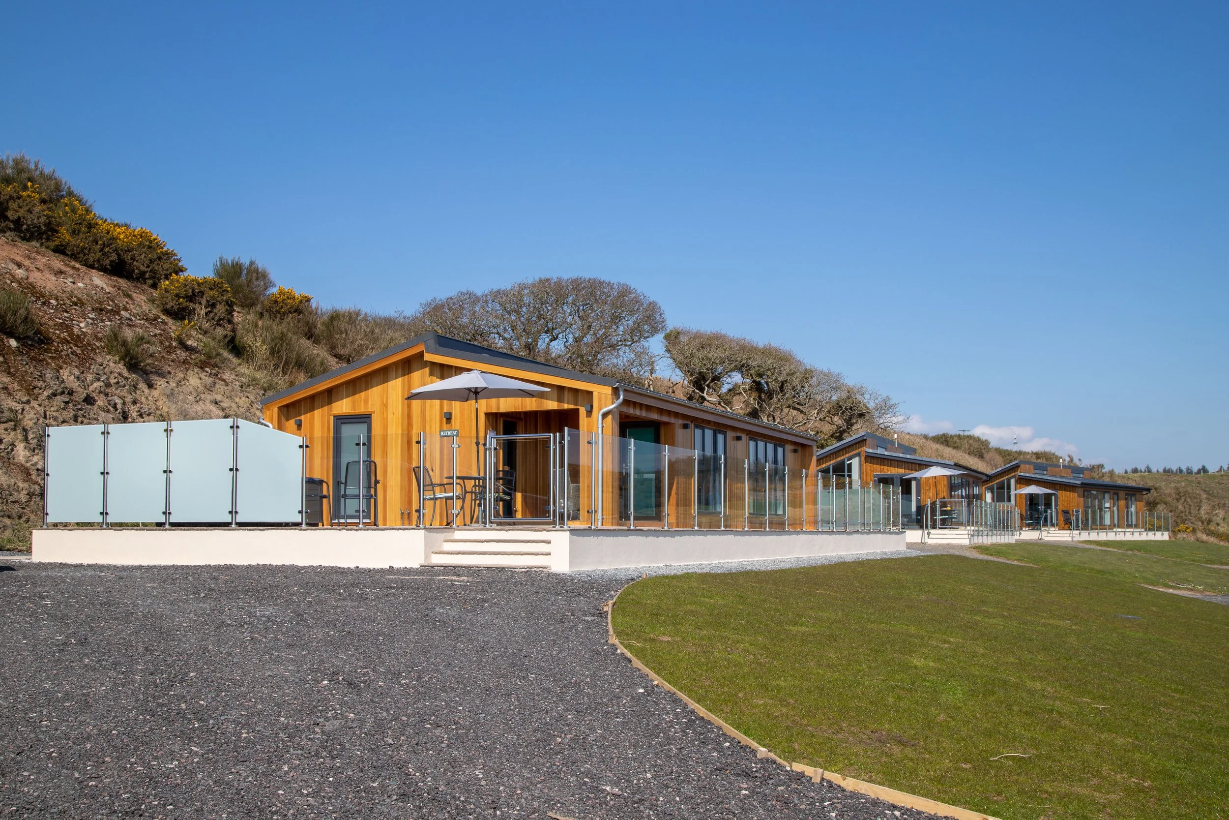 A row of modern wooden houses with glass balconies on a hillside, under a clear blue sky.