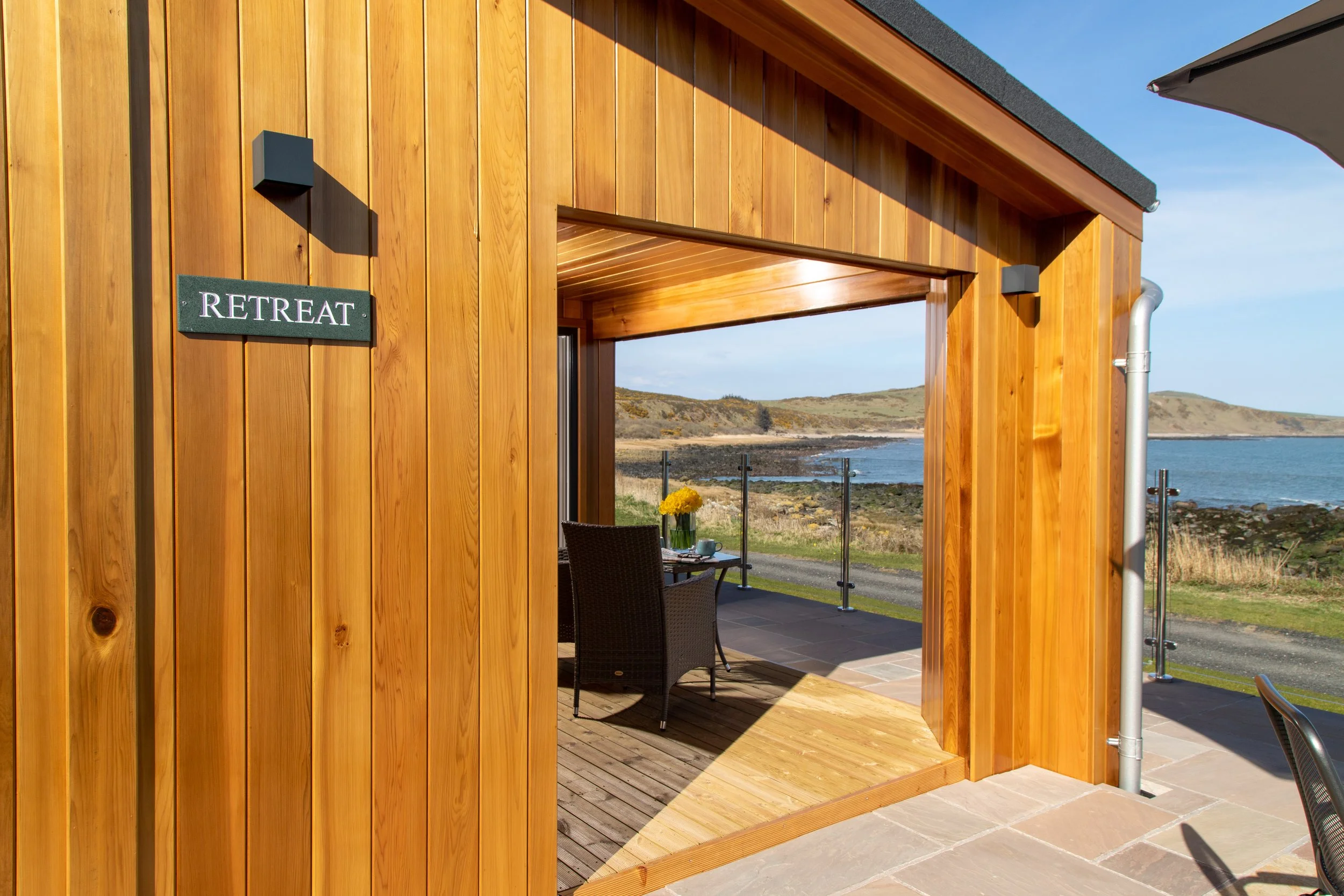 A wooden retreat overlooking a body of water with outdoor seating and a glass railing, under a clear blue sky.