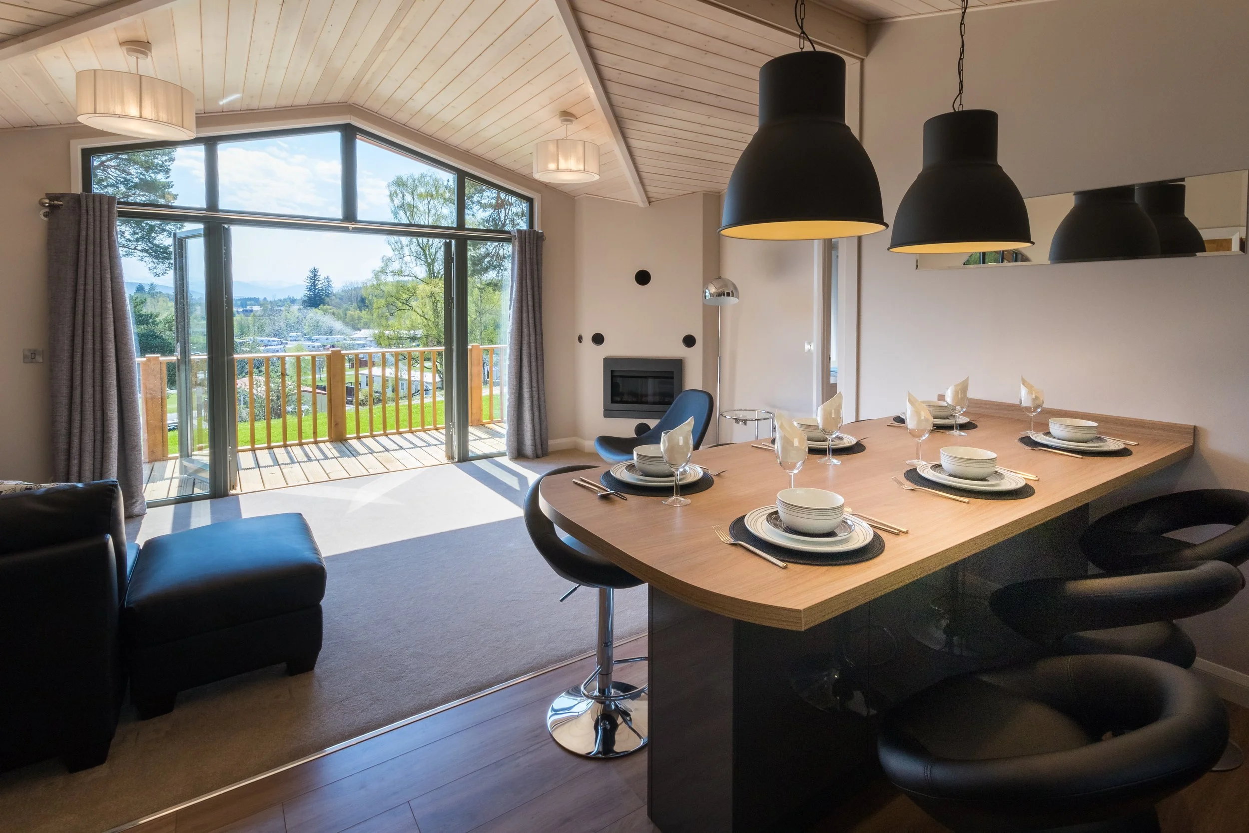 Modern living room and dining area with large windows, a balcony, black pendant lights, a wooden dining table set for six, and a view of trees and a parking lot outside.