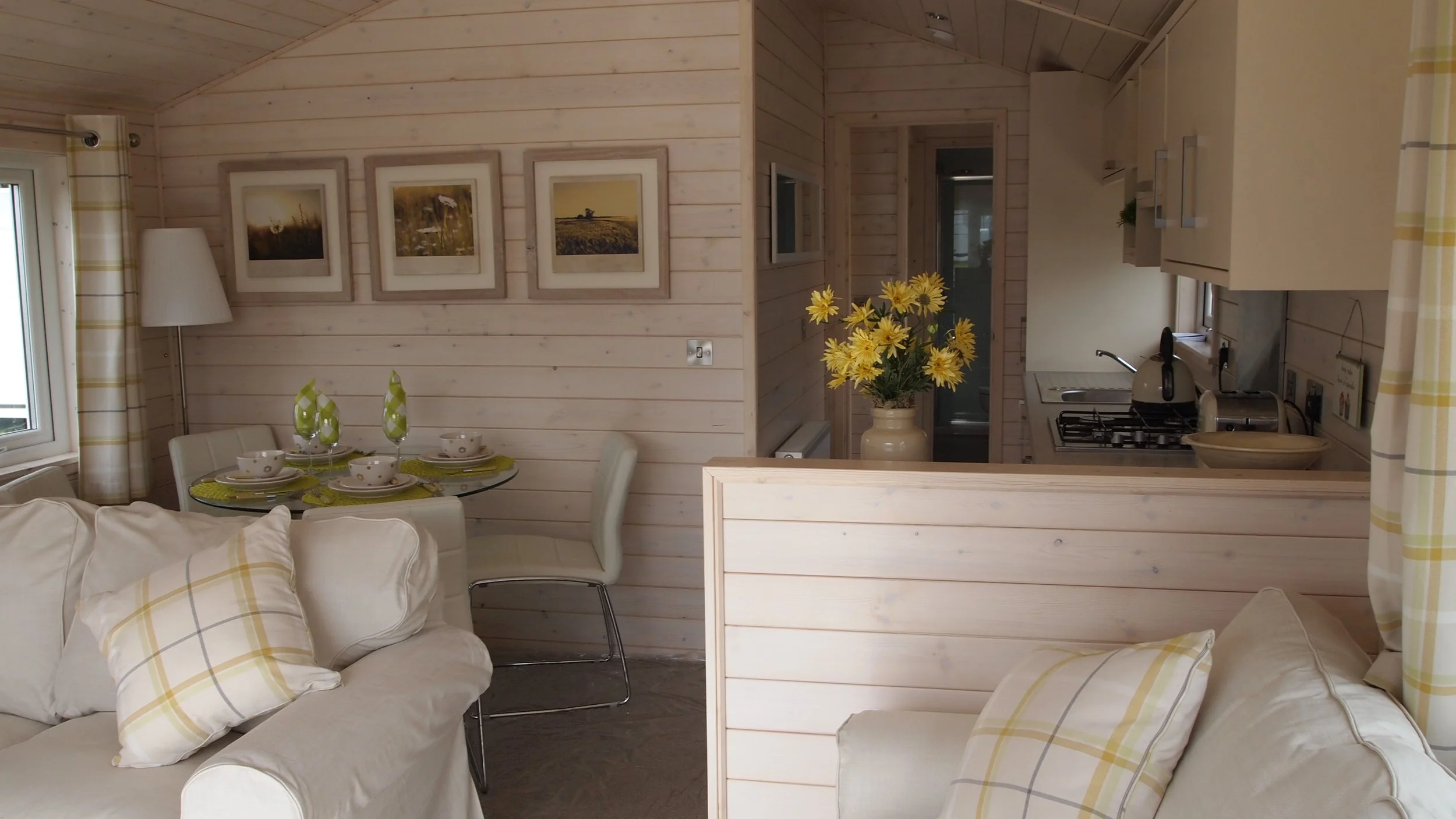 Living room and kitchen area with white furniture and wood-paneled walls, decorated with framed photos, a vase of yellow flowers on the kitchen counter, and a round dining table set with green and white tableware.