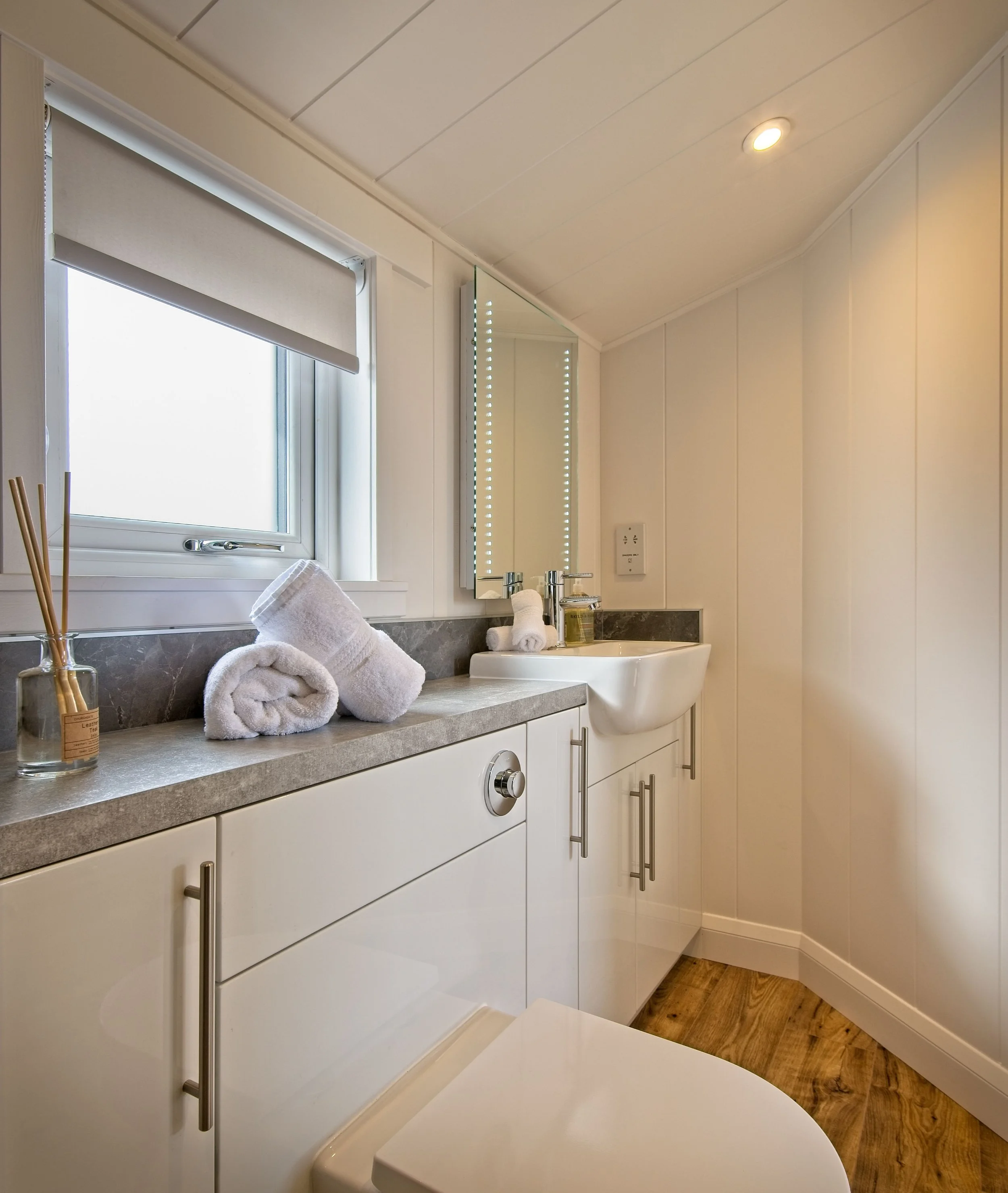 A modern bathroom with a white vanity, rolled towels, small toiletries, a window with a shade, a mirror, a sink, and wood flooring.