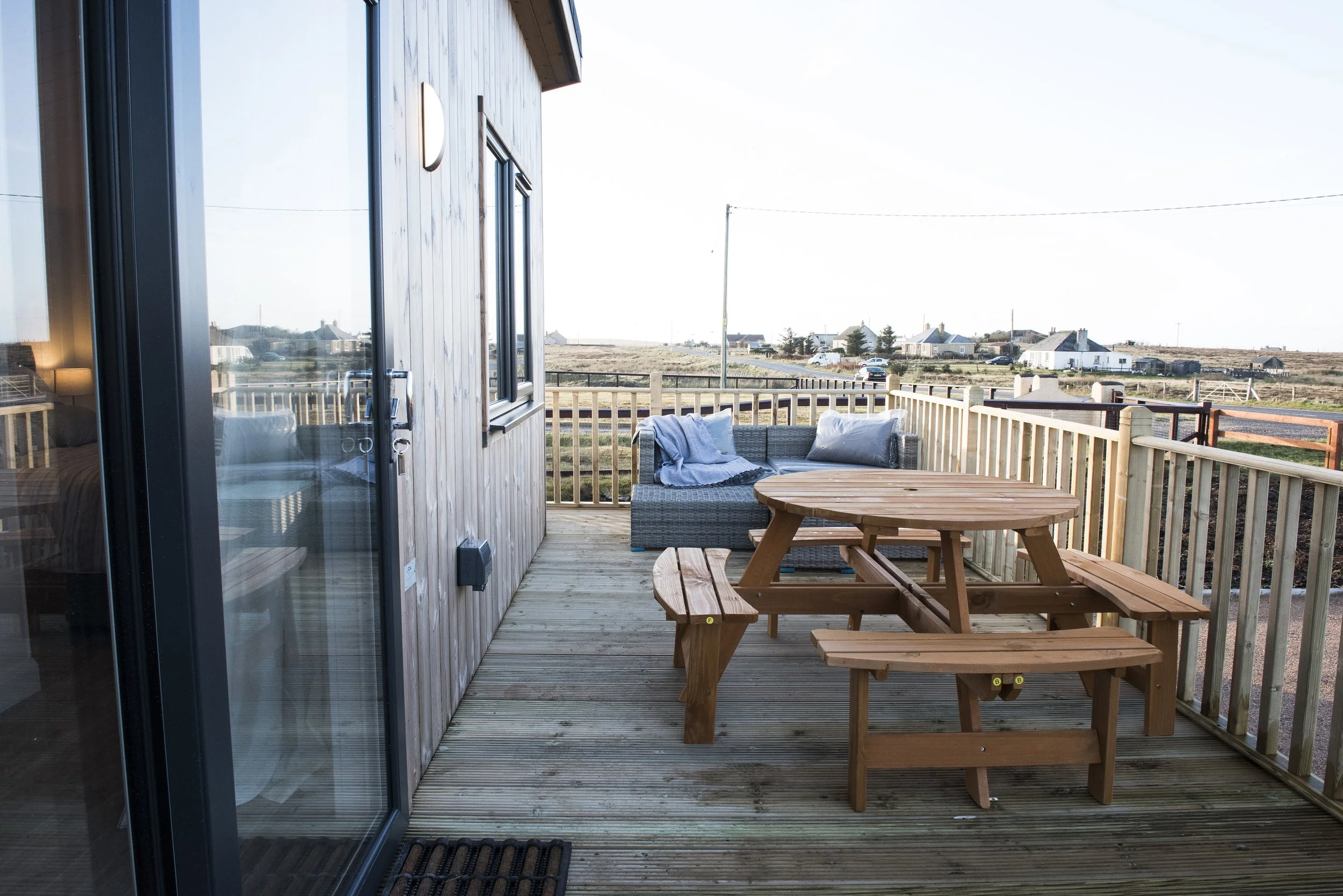 Wooden deck with picnic table, wicker sofa with cushions and throw blanket, and a view of rural landscape with houses and open fields.