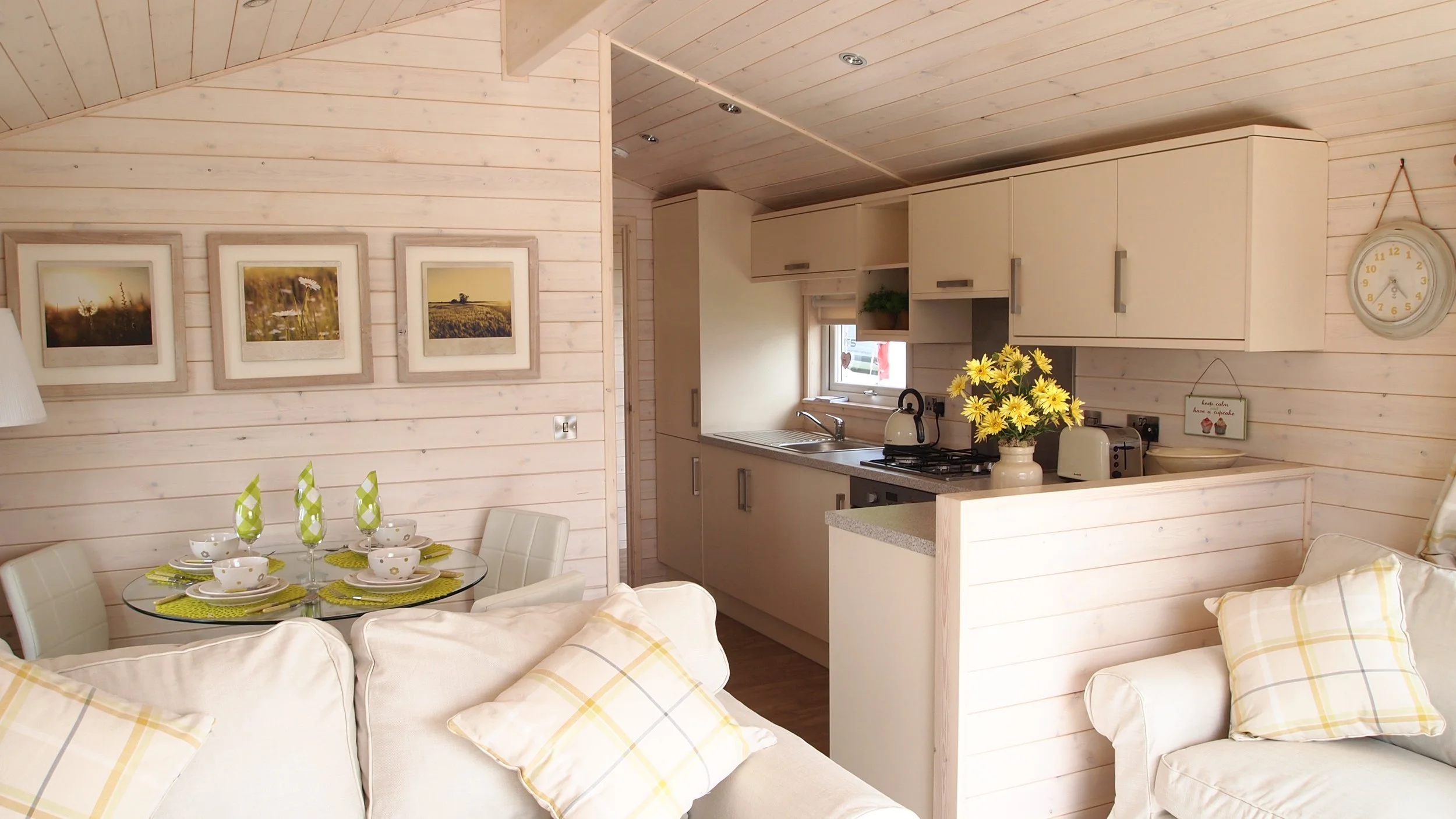 A cozy kitchen and dining area with cream-colored sofas, plaid pillows, a round glass dining table set with four place settings, and pastel-colored napkins. The kitchen has beige upper cabinets, a countertop with a vase of yellow flowers, a kettle, t