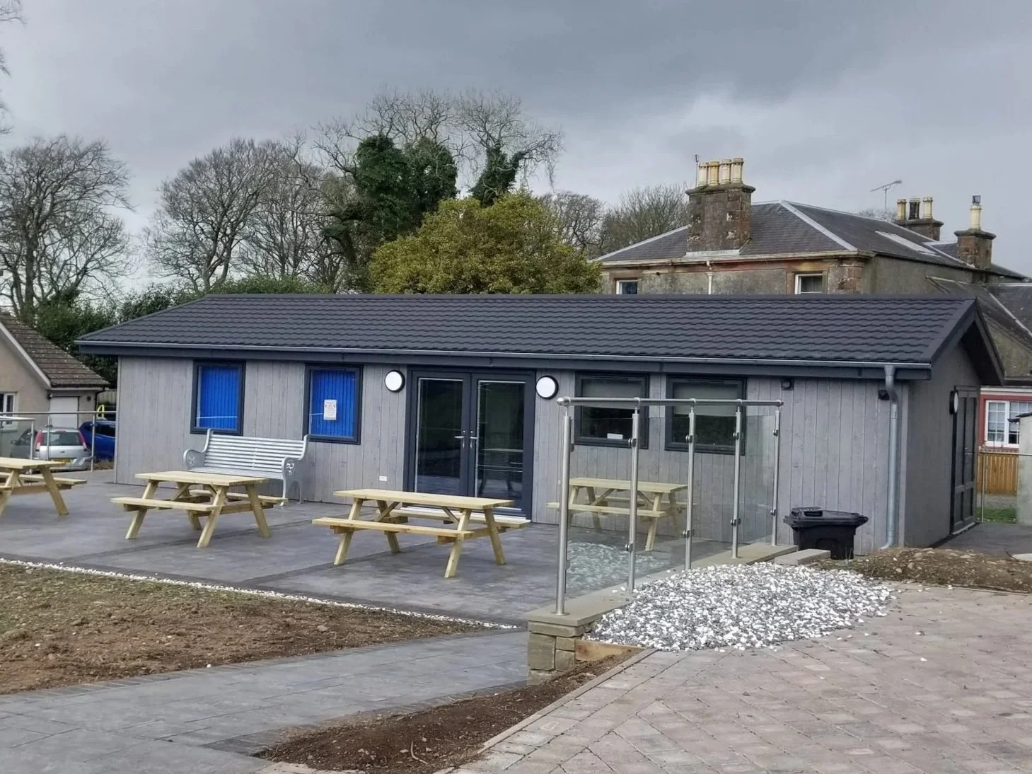 A modern building with gray wooden siding, a dark tiled roof, and three blue doors and windows, surrounded by outdoor seating and picnic tables, with a patio area and some landscaping under cloudy skies.
