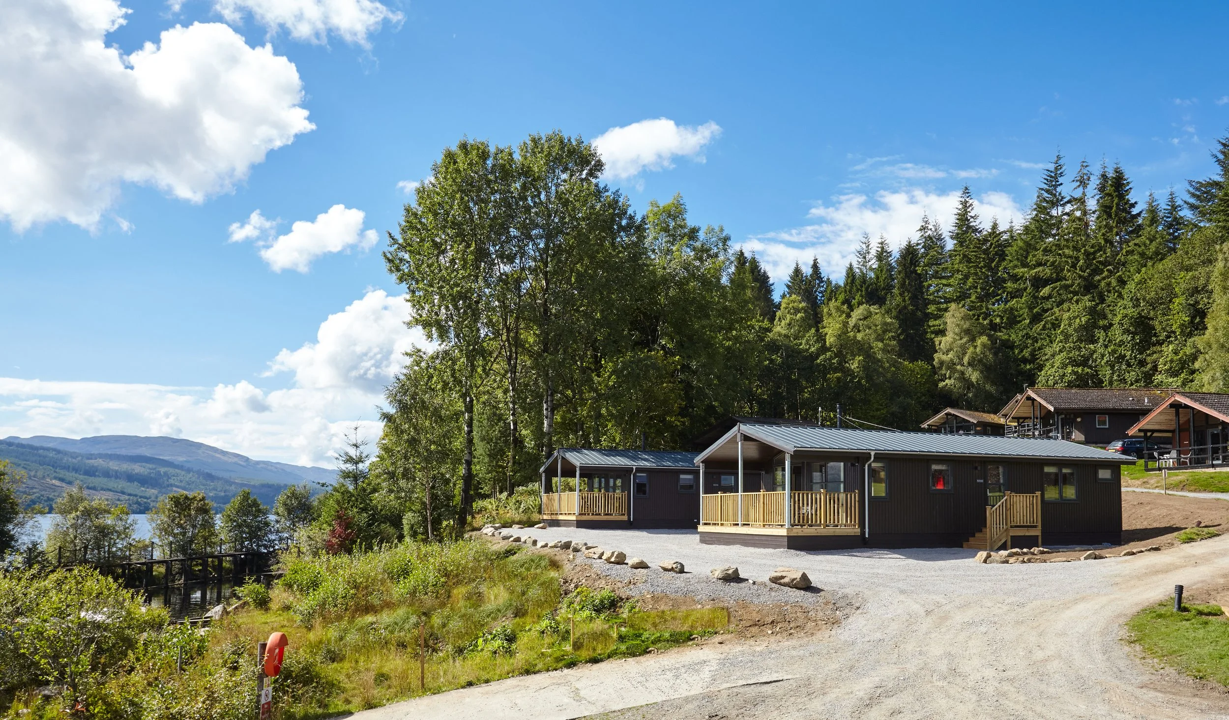 A cabin with a metal roof and wooden deck overlooking a scenic lake and forested hills on a bright, partly cloudy day.