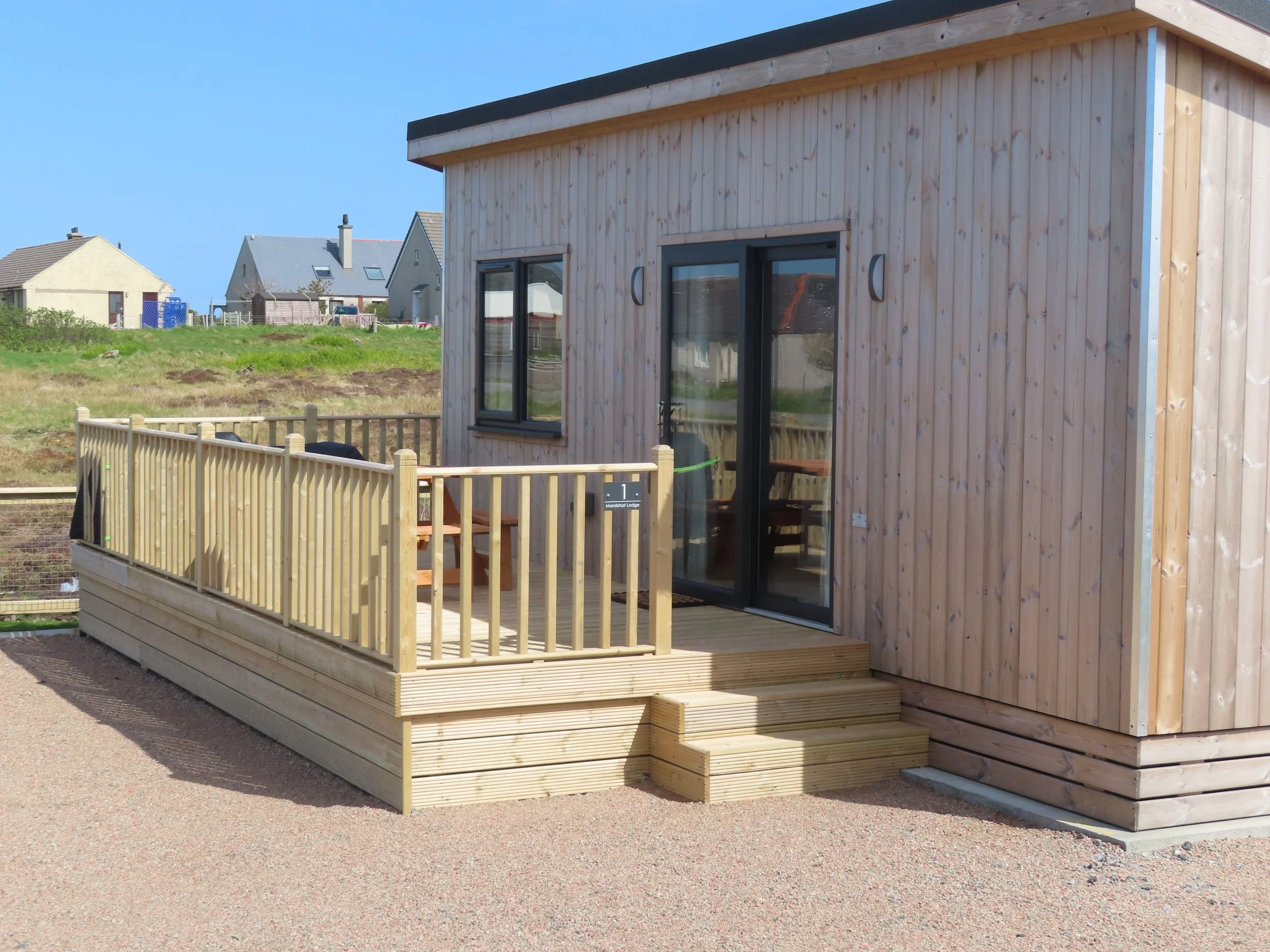 A small wooden house with a newly built deck and staircase outside, with sliding glass doors and windows, set in a suburban area with other houses in the background.