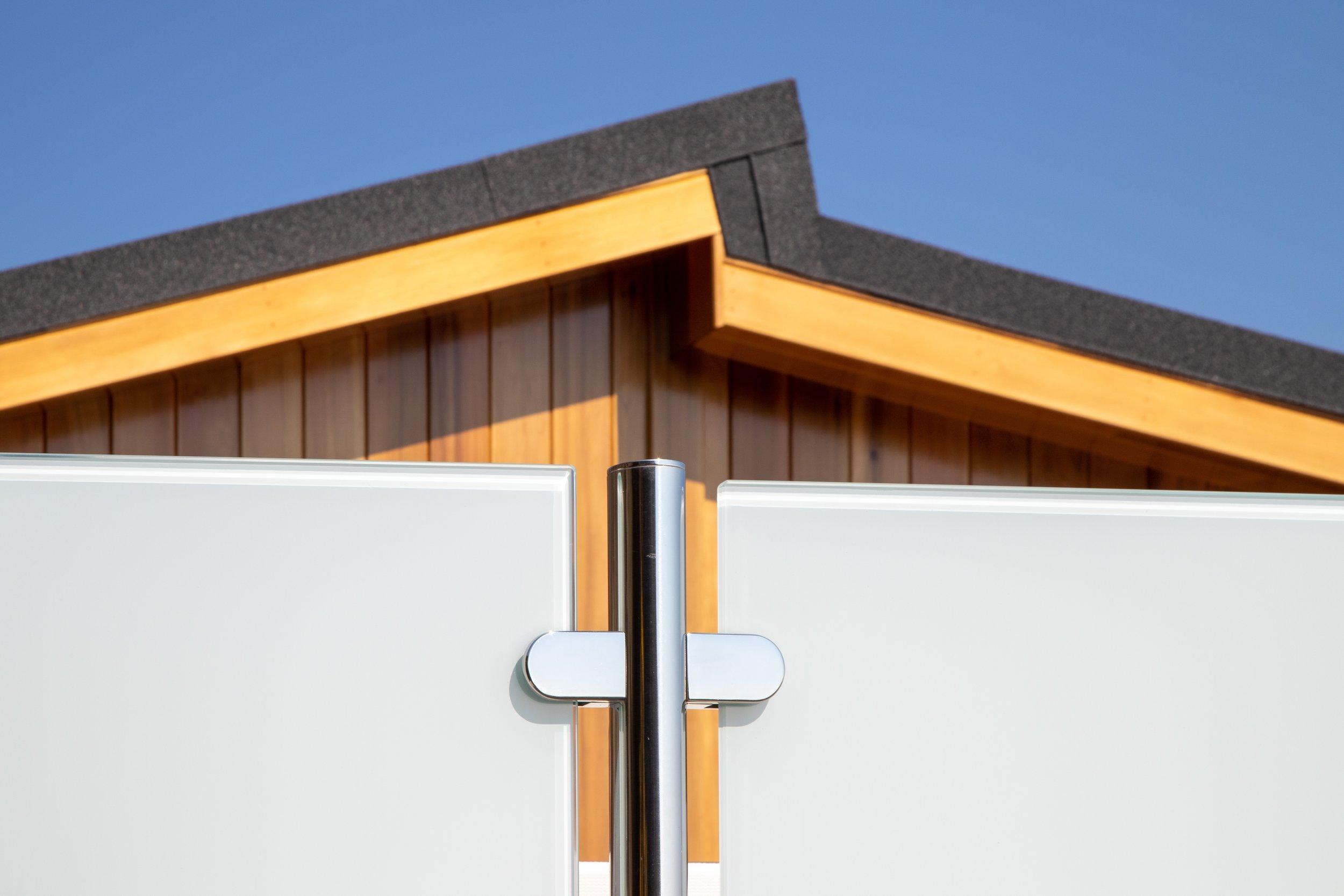 Close-up view of a white outdoor privacy screen with a metal hinge, with a wooden house with a sloped roof in the background under a clear blue sky.