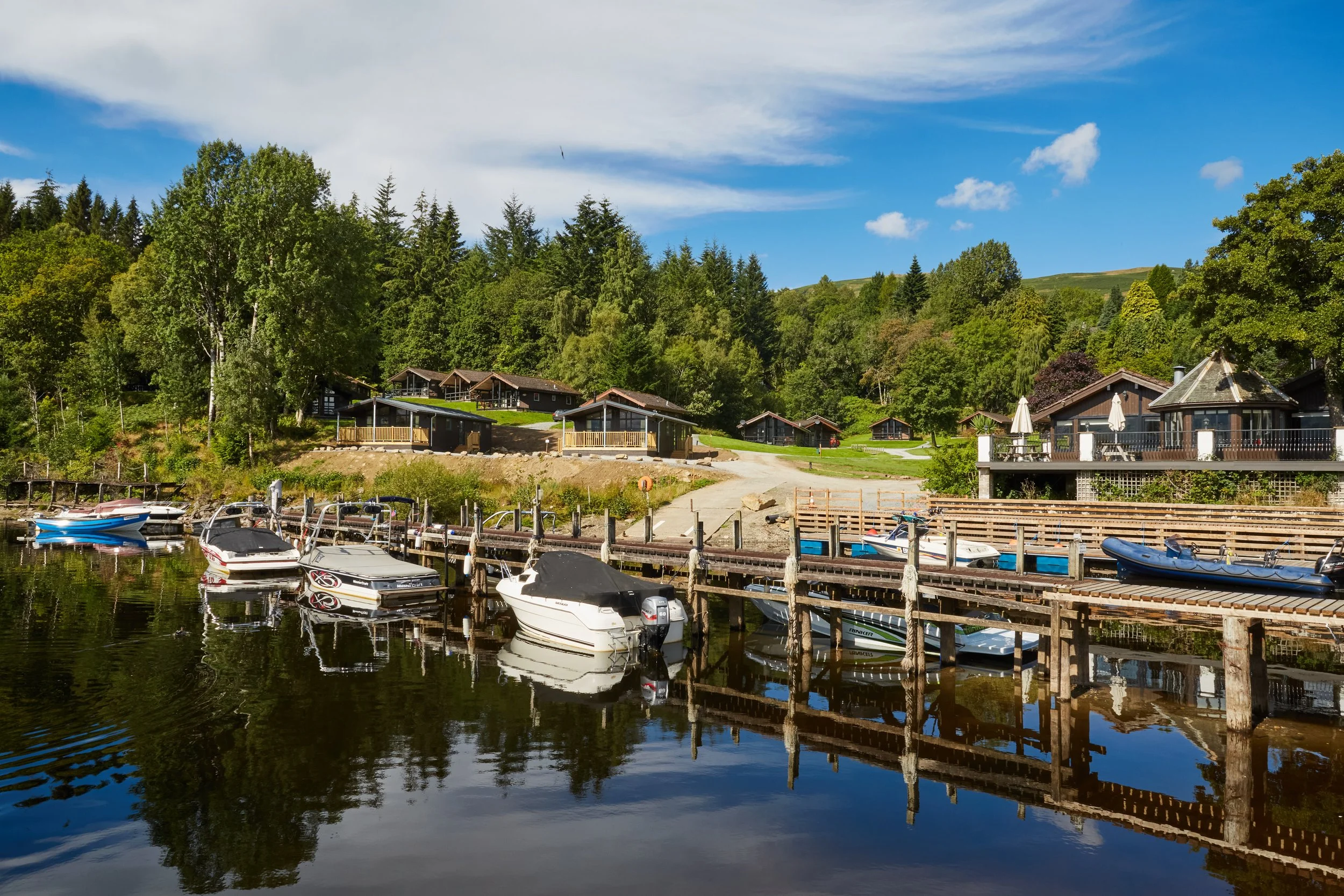 Boats docked at a marina with wooden piers near waterfront houses surrounded by lush green trees and a hillside in the background under a partly cloudy sky.