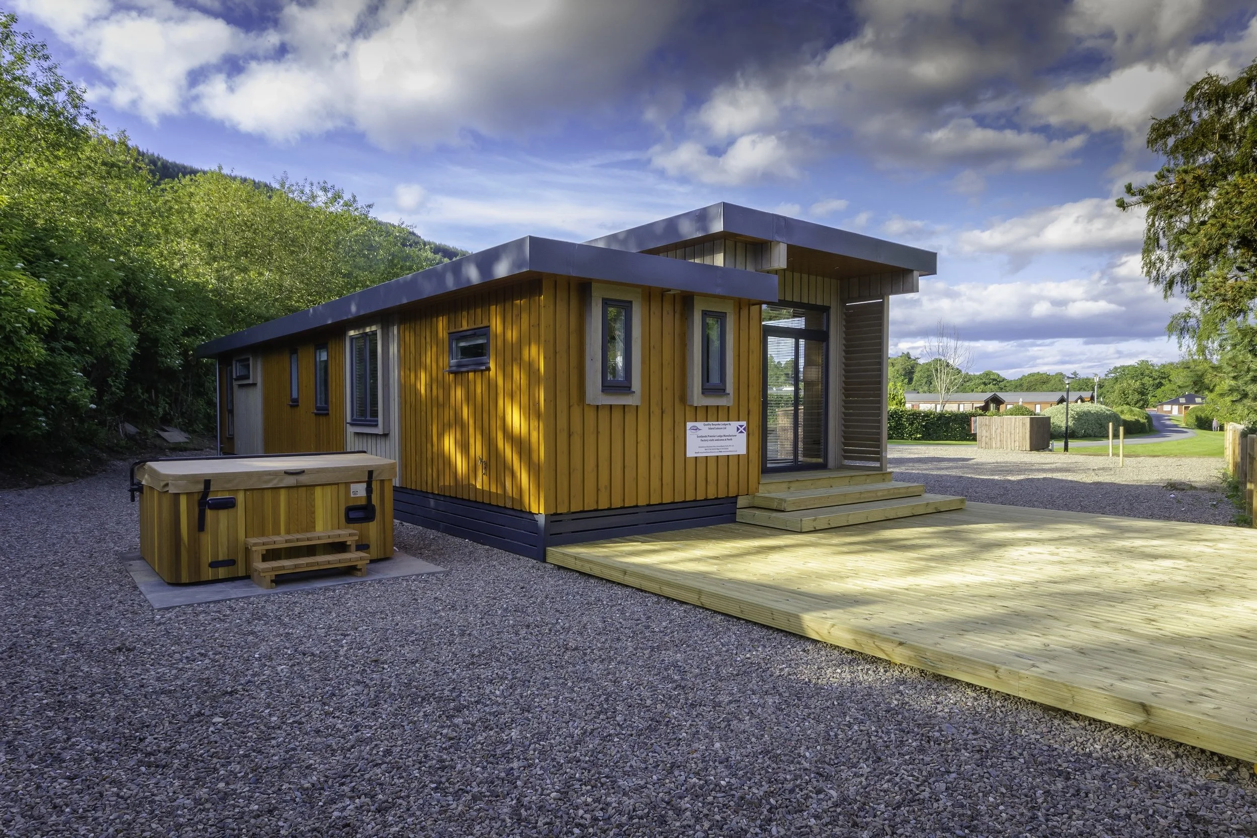 A small modern wooden house with steps leading to the entrance, situated on a gravel surface, with a hot tub outside, surrounded by green trees and a partly cloudy sky.