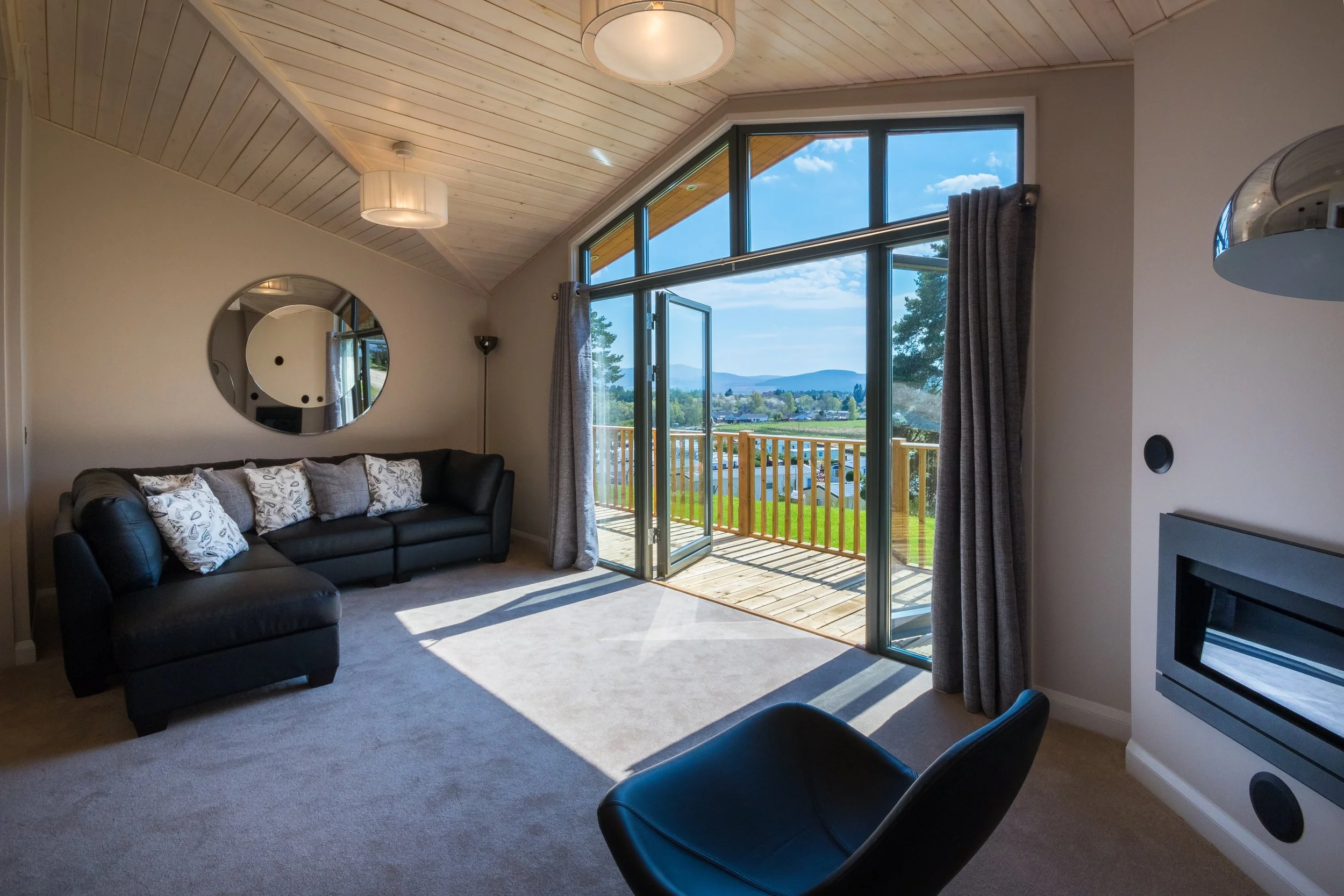 Living room with large glass doors opening to a balcony with scenic mountain view, black sofa with decorative pillows, circular mirror on wall, wood-paneled ceiling, modern lighting fixtures, and carpeted floor.
