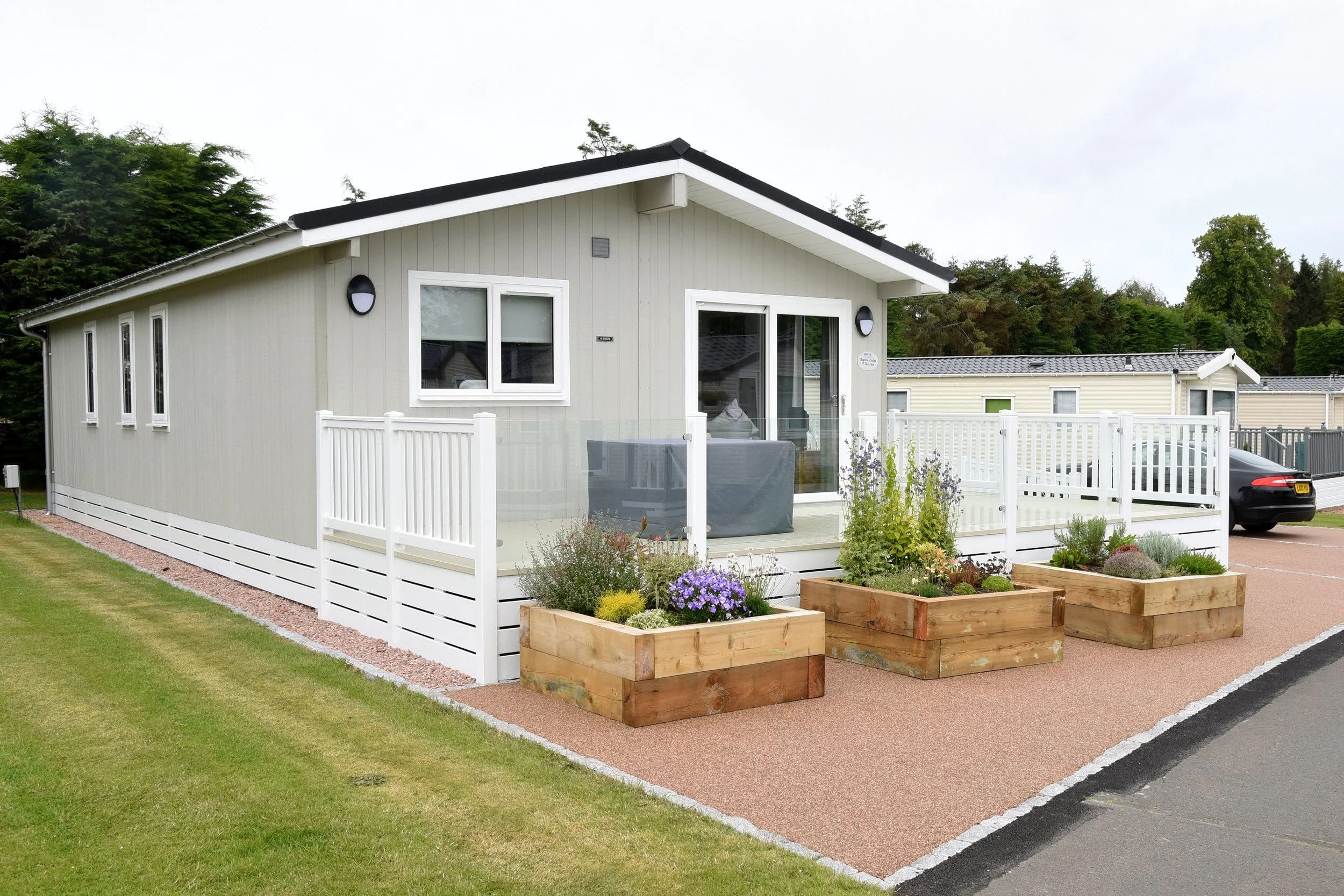 A small, modern, single-story house with light-colored siding and a gabled roof, surrounded by a well-maintained lawn, flower beds, and a white picket fence on a porch with outdoor furniture.