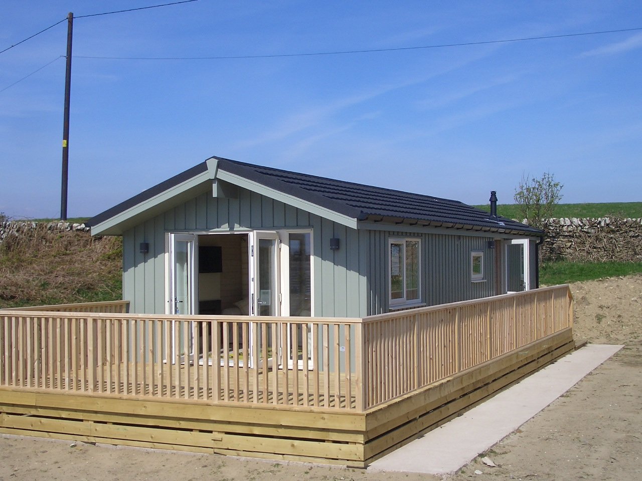 A small modern house with a gray exterior, black roof, and a wooden deck with a railing, situated on a dirt lot with a stone wall and green fields in the background.