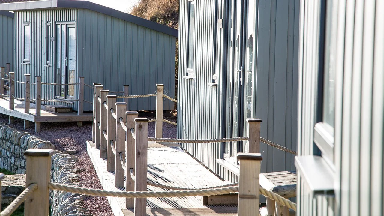 A wooden walkway with rope railing runs along the side of light-colored metal buildings with small windows.