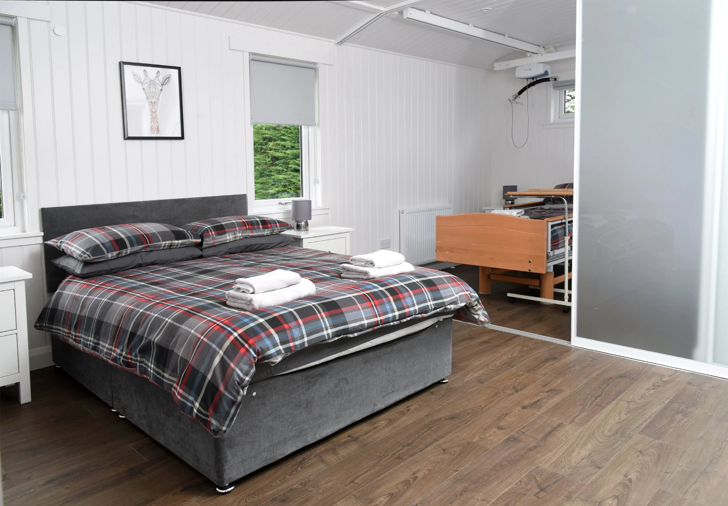 A bedroom with a bed covered in plaid bedding, white nightstands, a small lamp, a window with a view of green trees, and a hospital bed or recliner in the corner.
