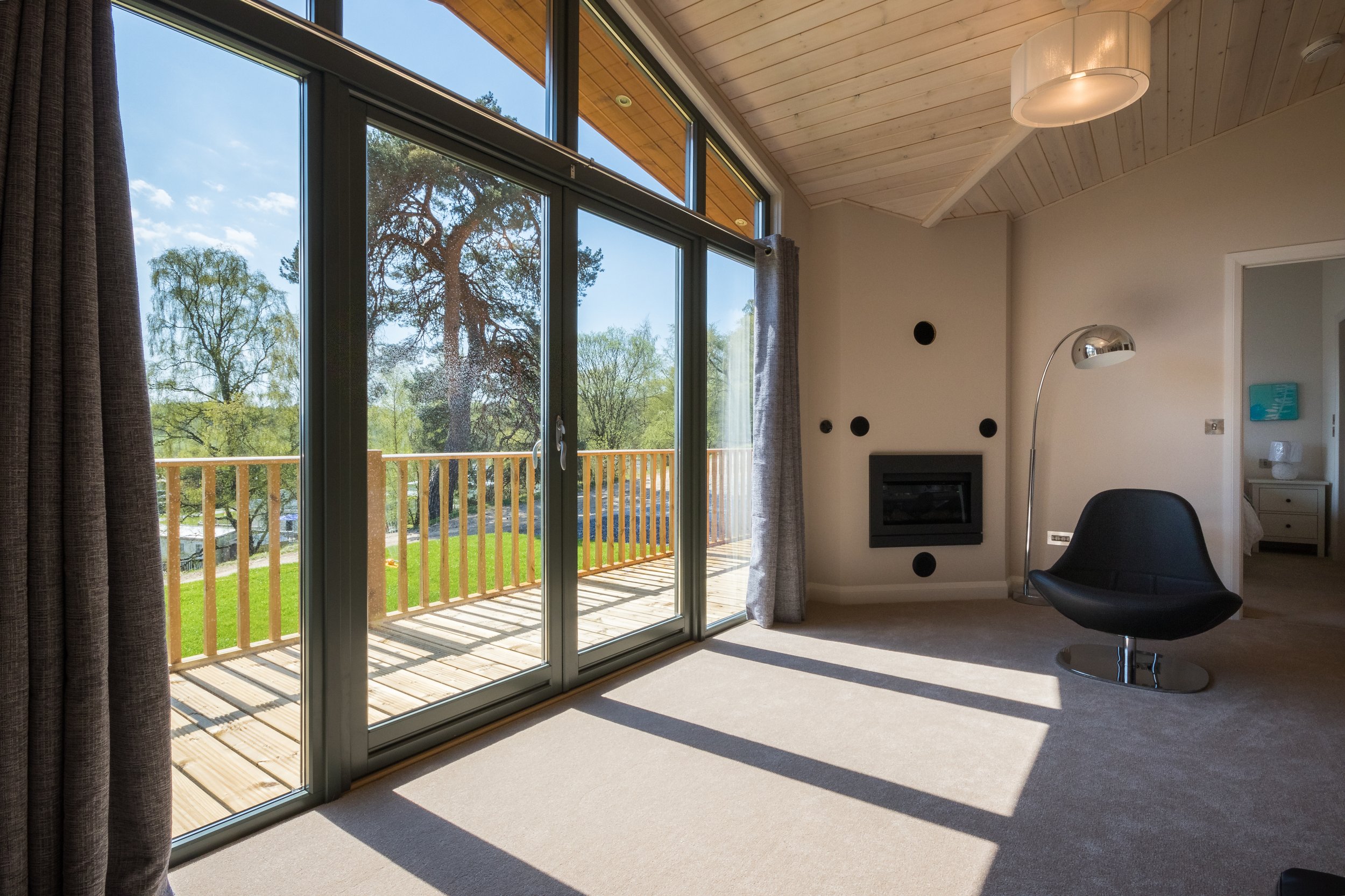 Living room with large glass sliding door leading to a balcony, beige carpet, wall with black dot decorations, black fireplace, and modern black lounge chair illuminated by sunlight.