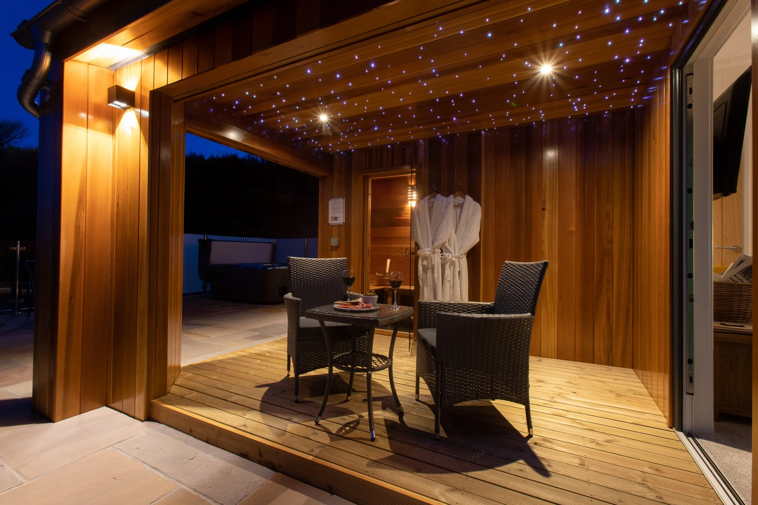 An outdoor wooden patio with string lights overhead, a small round table with four chairs, two wine glasses, a plate of food, and bathrobes hanging on the wall.